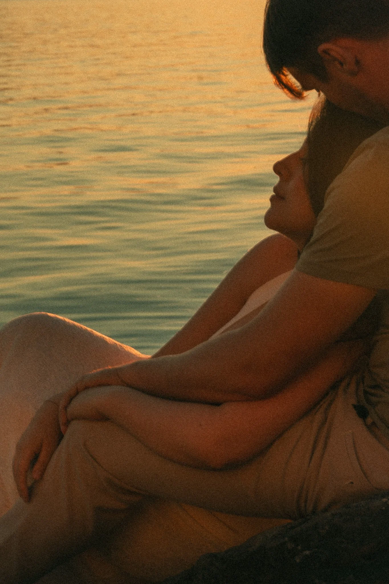 A couple sitting close by a body of water during sunset, with their faces touching and eyes closed.