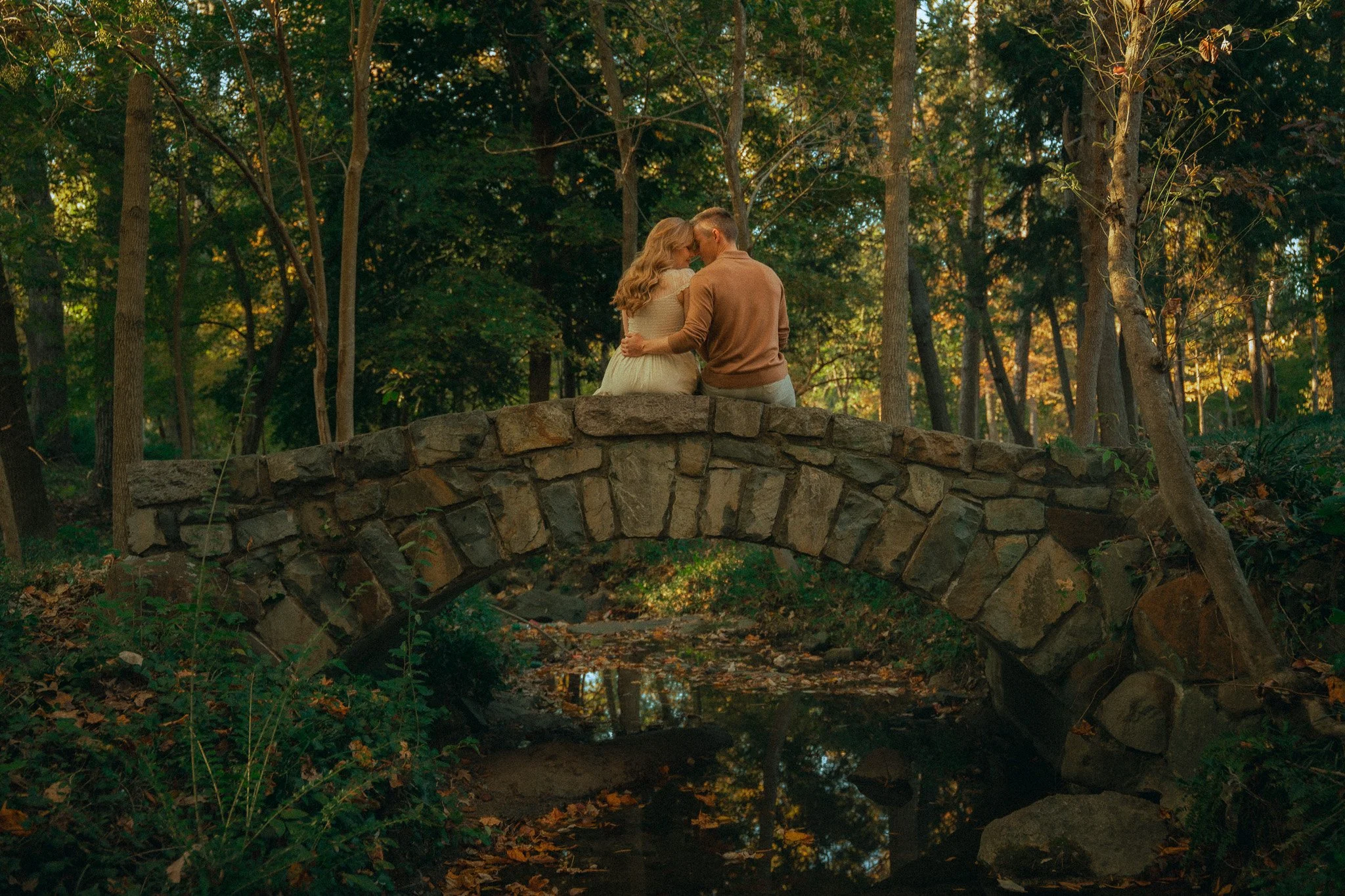 A couple sitting on a stone bridge in a wooded park during autumn, embracing and touching foreheads, surrounded by trees with fall foliage.