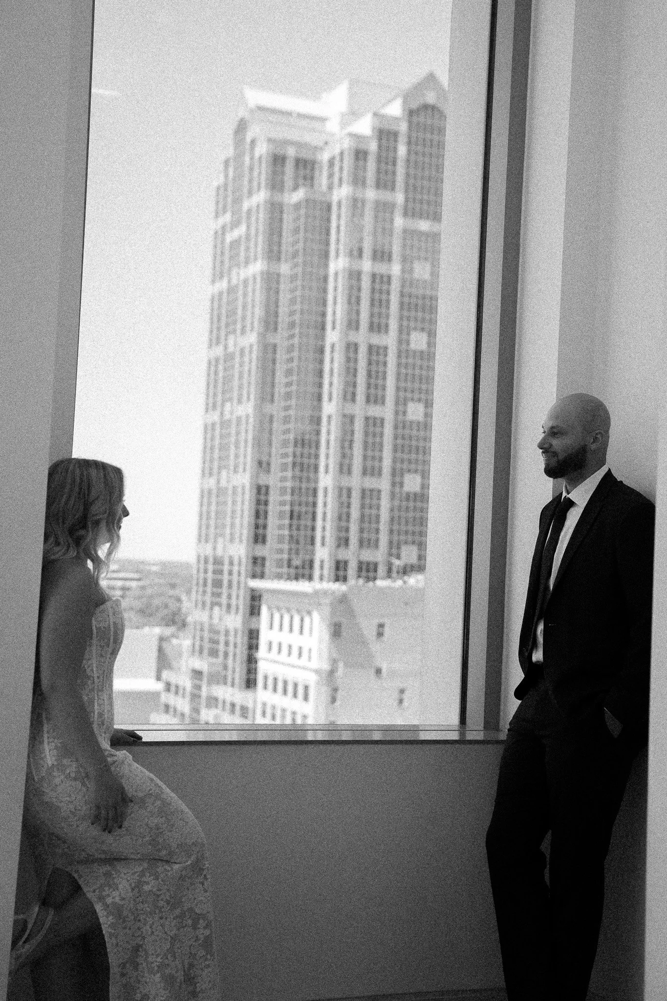 New York City engagement wedding documentary style photographer. A woman in a lace dress sitting on a ledge by a large window, and a man in a suit standing inside a tall building, with a cityscape of skyscrapers outside.