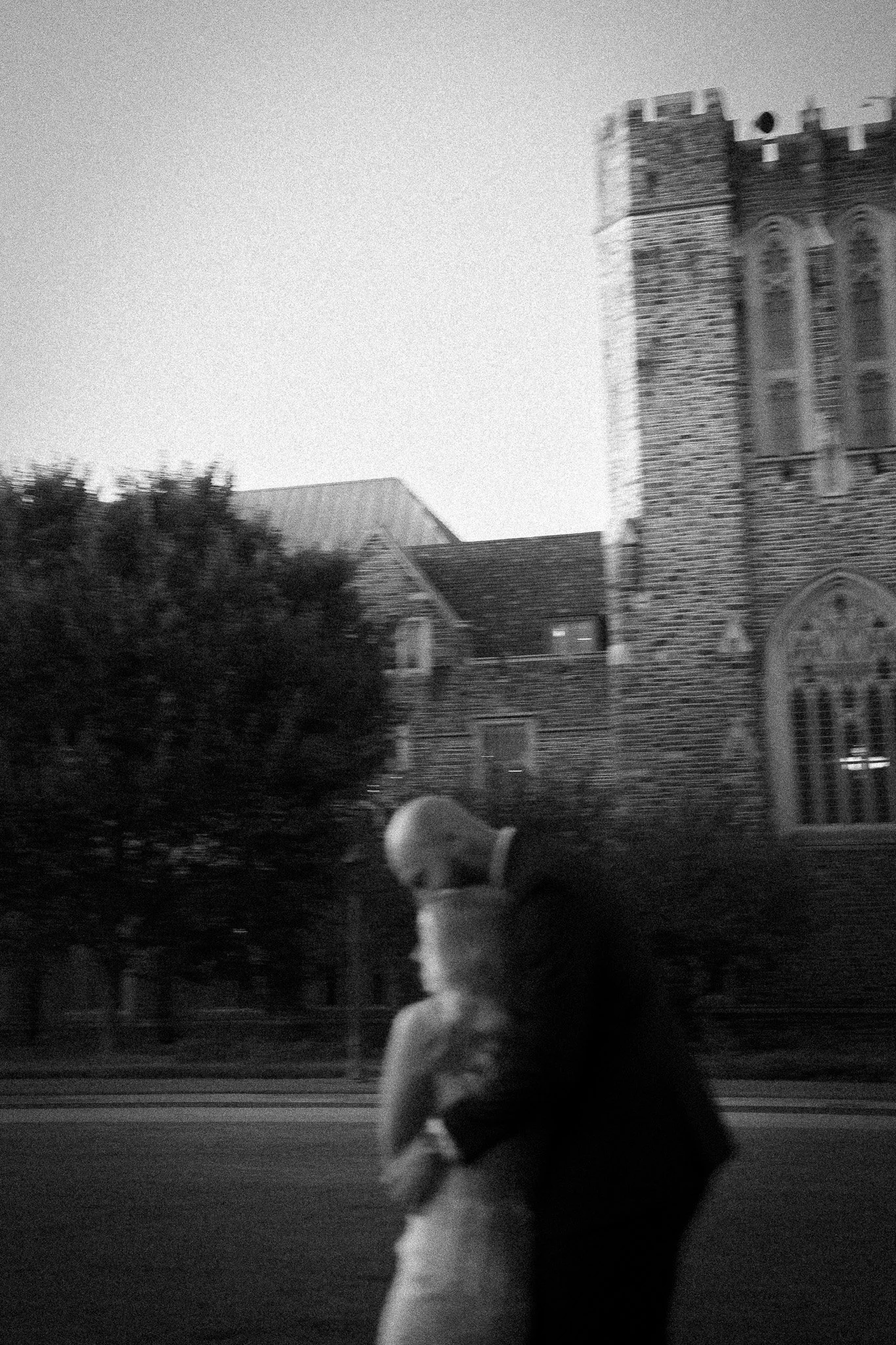 A black and white photo of a man kissing a woman, with a brick church and trees in the background.