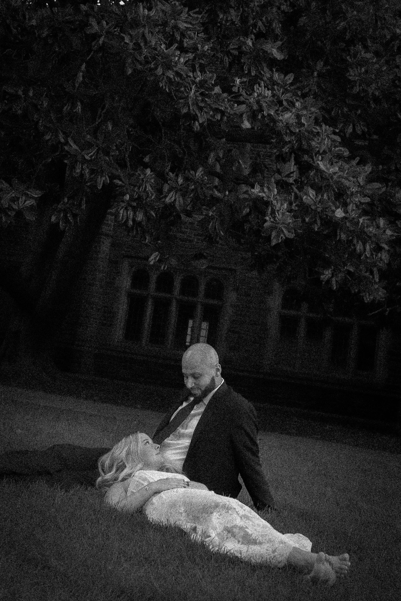 A black-and-white photo of a man and a woman outdoors at night. The woman is lying on the grass, looking up at the man, who is sitting beside her and looking down at her. There are large trees and an old building in the background.