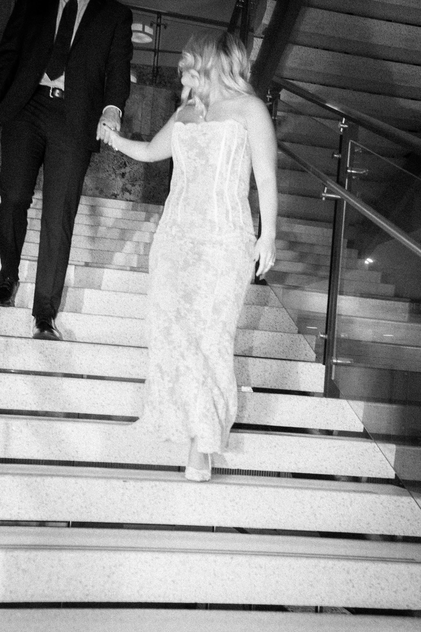 Editorial Pinterest board photographer. A woman in a long lace dress descending stairs while holding hands with a man in a suit, in an indoor setting.