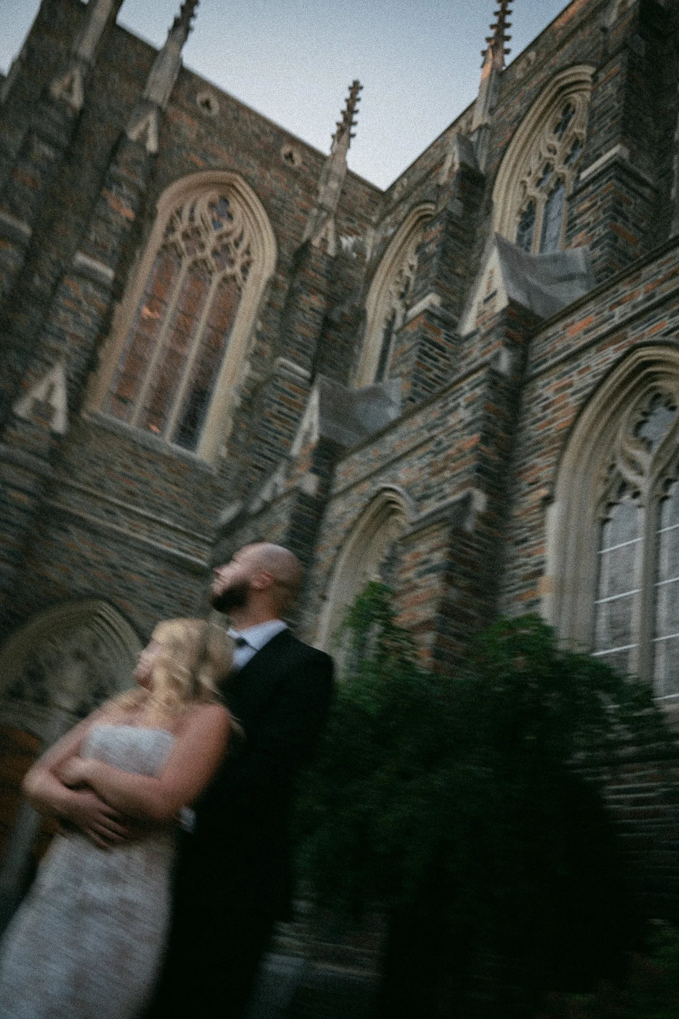 A couple dressed in wedding attire embracing outside a historic brick church with tall, narrow windows and decorative stone detailing.