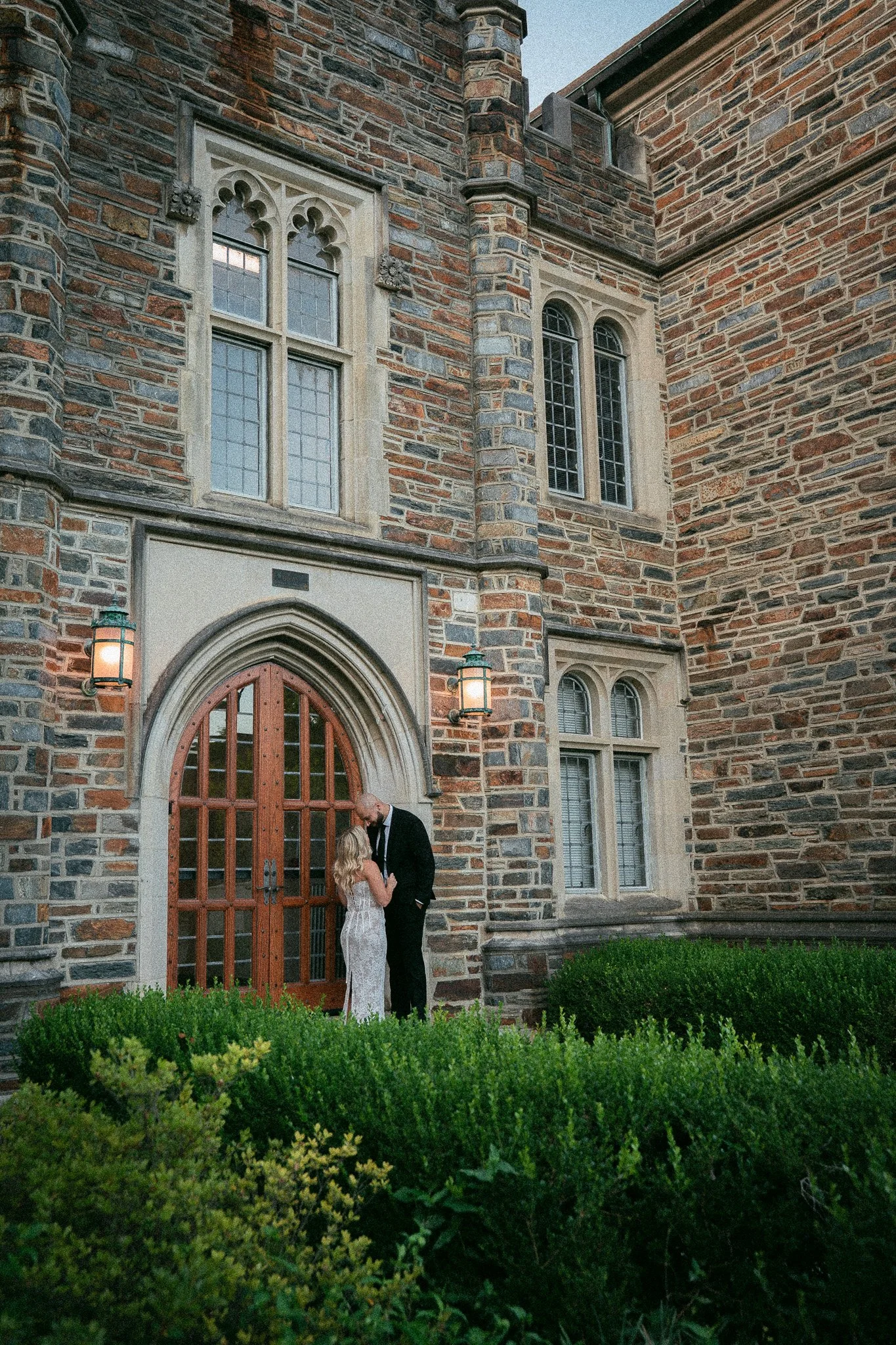 A couple dressed in formal attire standing close together outside a large brick building with gothic-style windows and a wooden arched door, surrounded by green bushes.