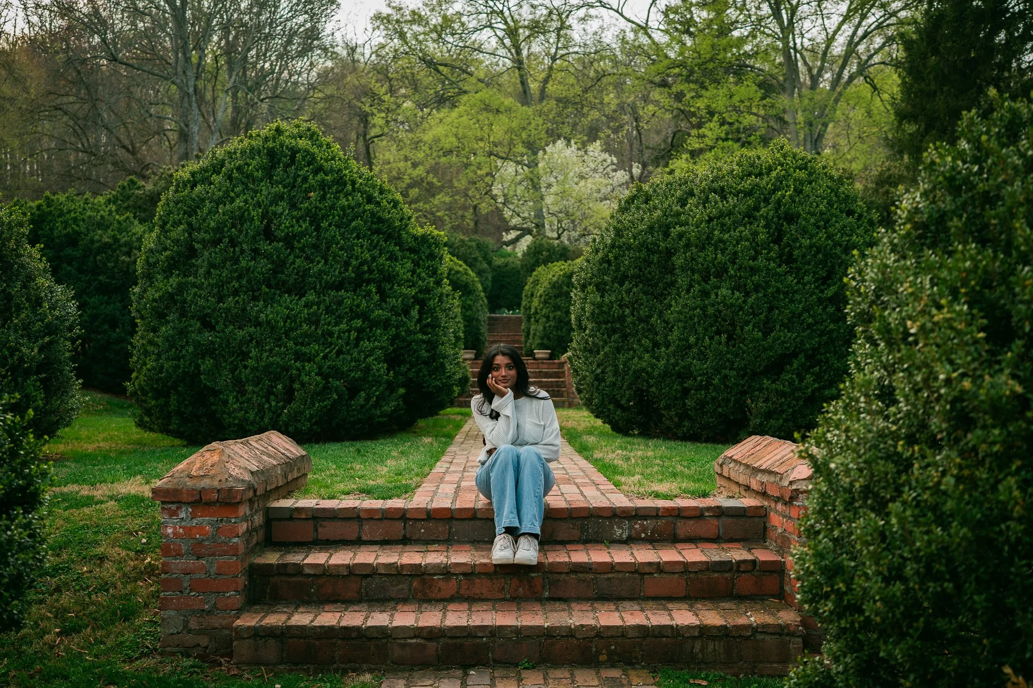 A woman in a white top and light blue jeans sitting on brick steps in a garden with lush green bushes and trees in the background.