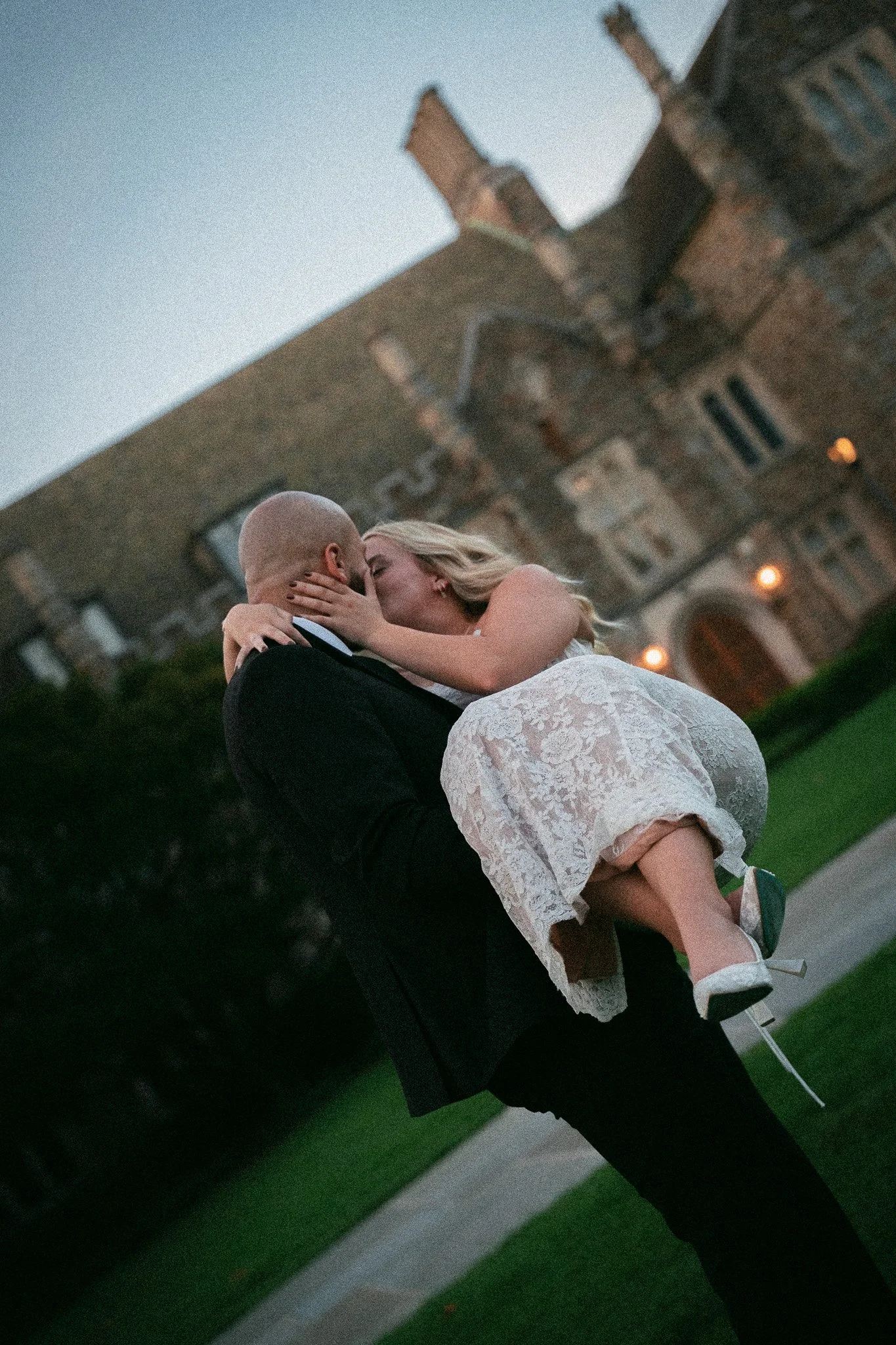 A man in a black suit carrying a woman in a white lace dress and white high heels, sharing a kiss outside a large brick house during dusk.