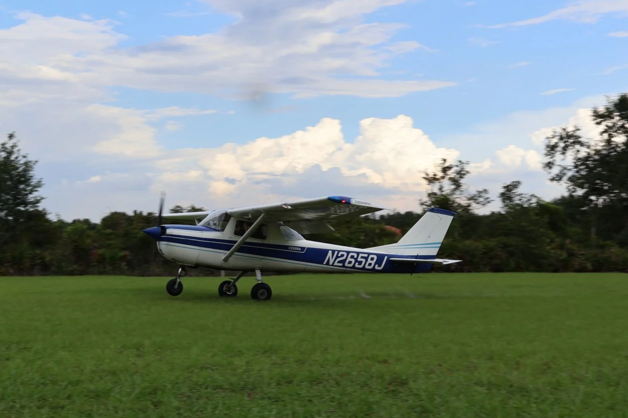 Small white and green single-engine airplane with gold stripes parked on a grassy field under a partly cloudy sky.