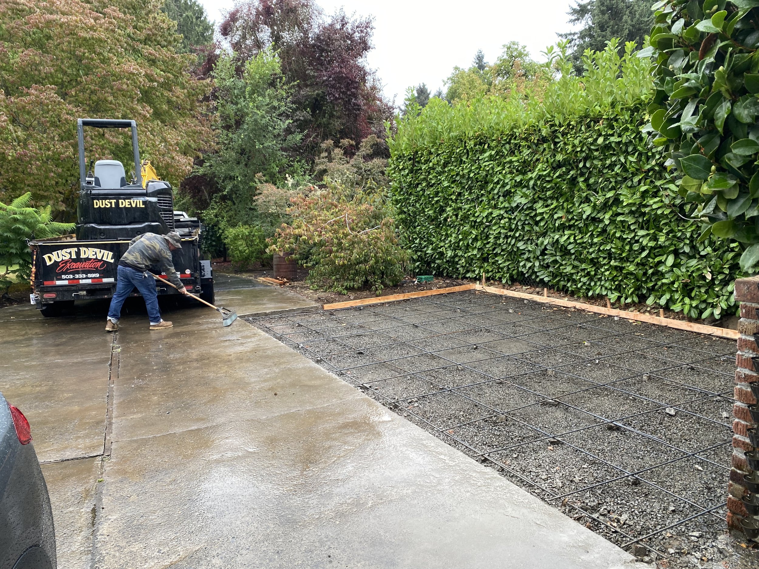 A worker is leveling wet concrete on a driveway with a long-handled tool, with a green hedge and trees in the background, and a black pickup truck labeled 'Dust Devil Excavation' nearby.