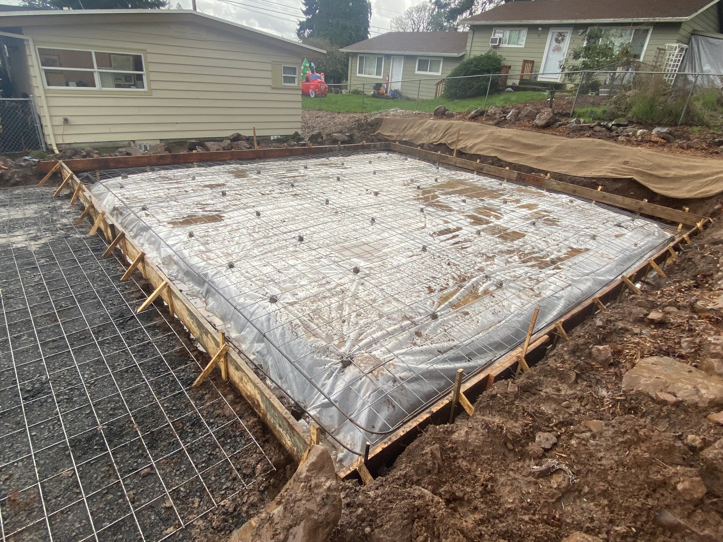 Construction site with rebar reinforcement prepared for pouring concrete, with sandbags lining the edges and residential houses in the background.