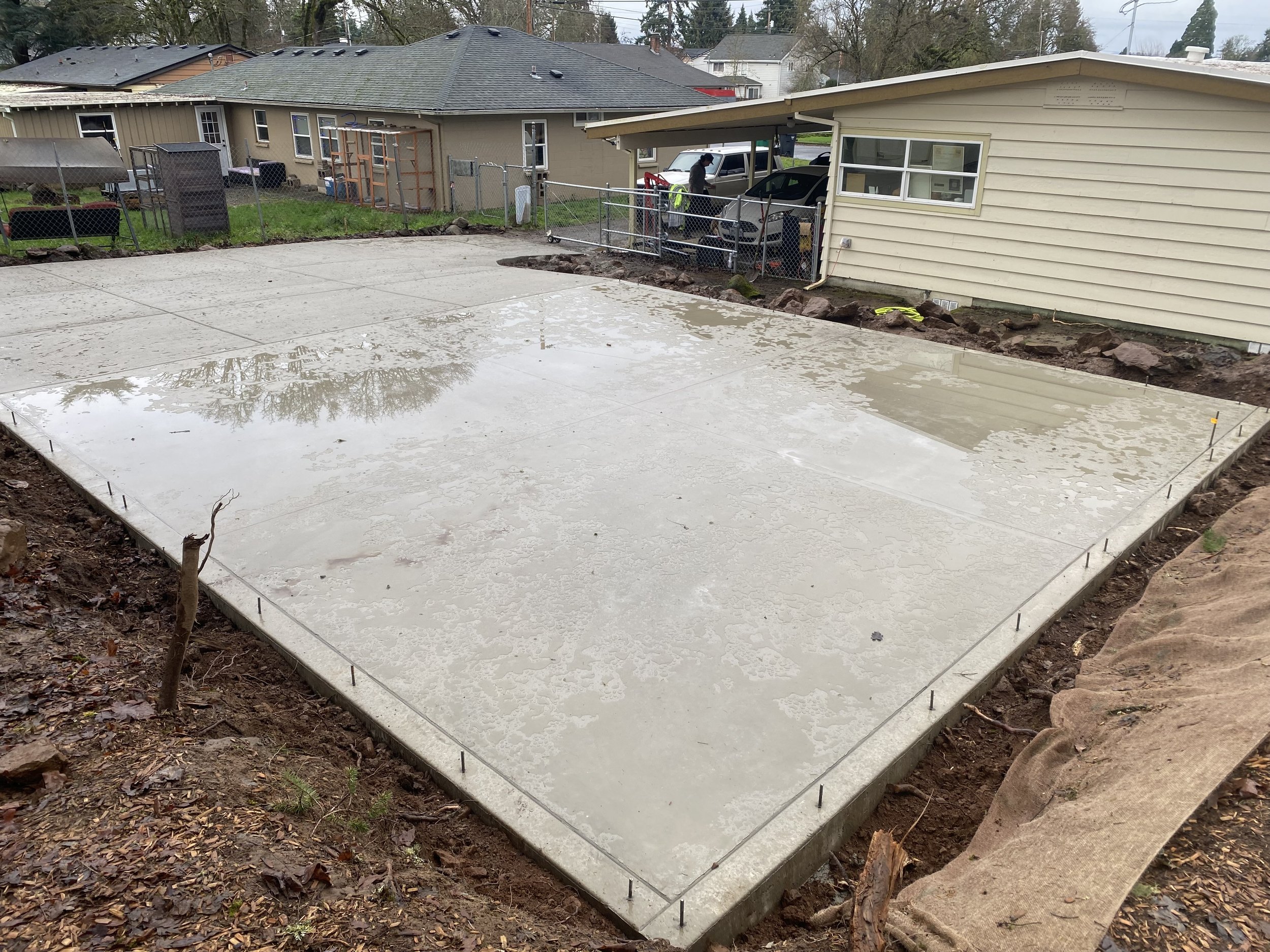 Concrete foundation poured in a backyard for a construction project, with an adjacent house, ladder, and construction worker in the background.