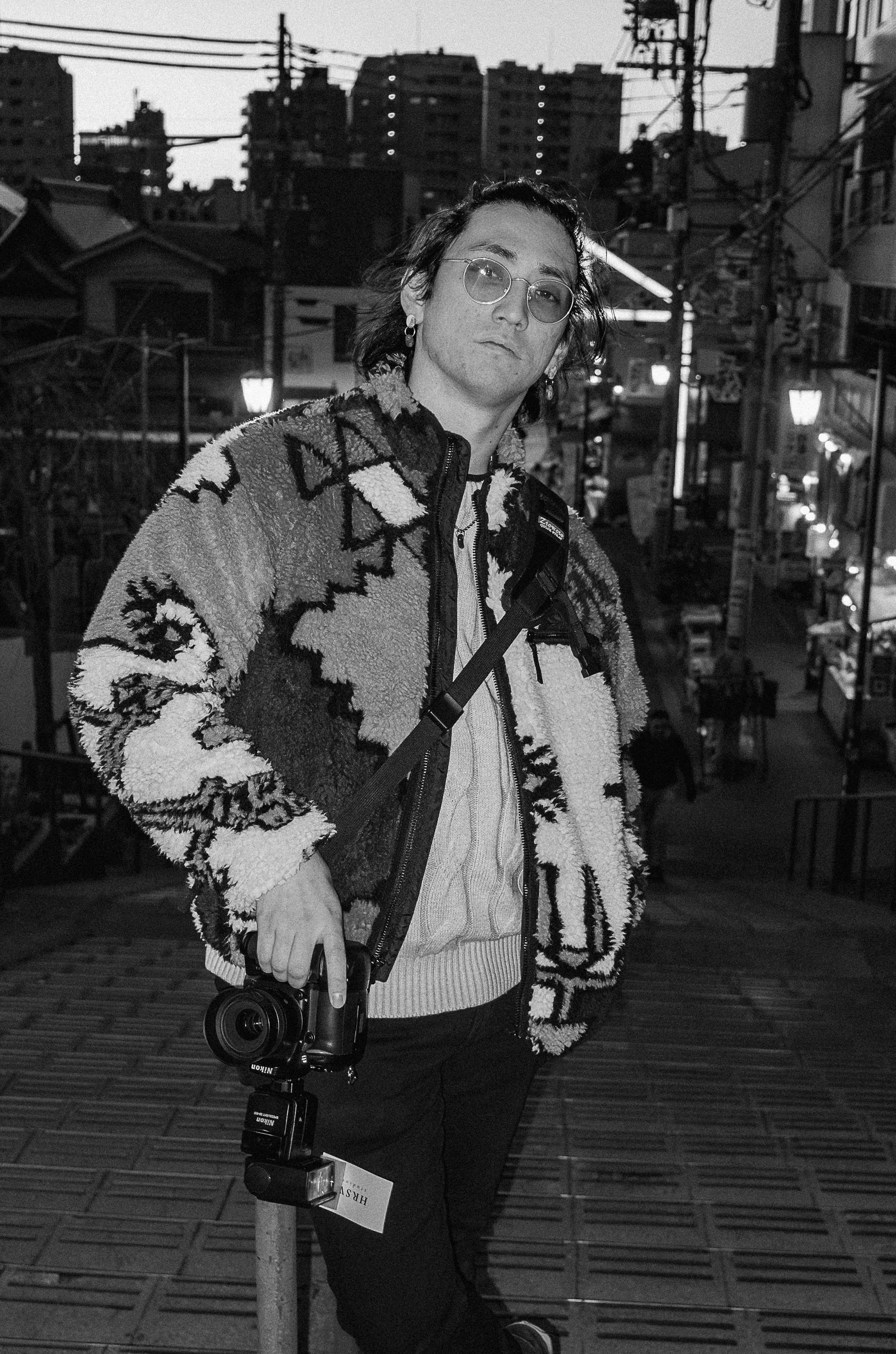 A person with shoulder-length hair, glasses, and earrings standing on a city street at dusk, holding a camera in their right hand and wearing a patterned fleece jacket and cables around their neck.
