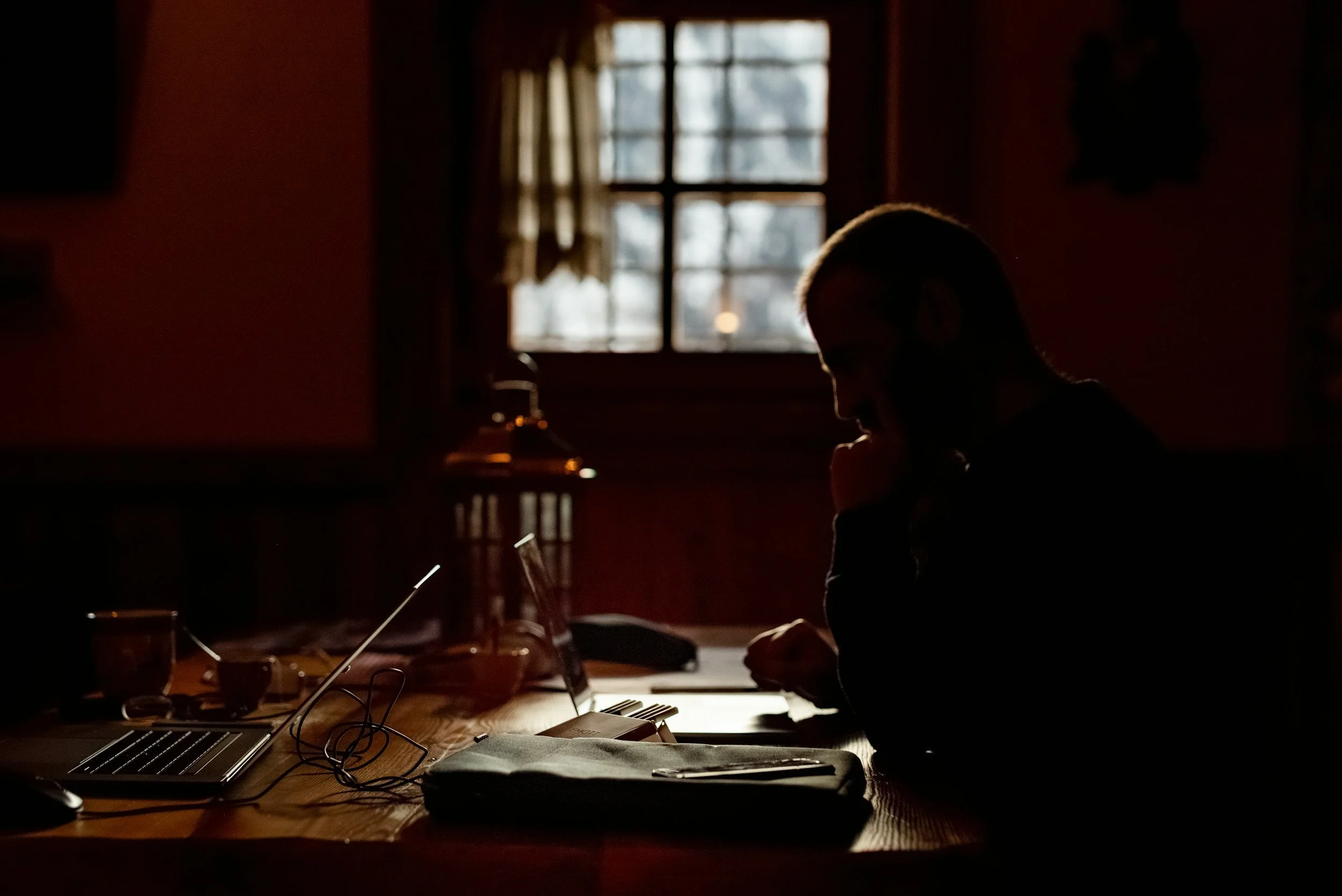 A silhouette of a man sitting at a desk in a dimly lit room with a window in the background.