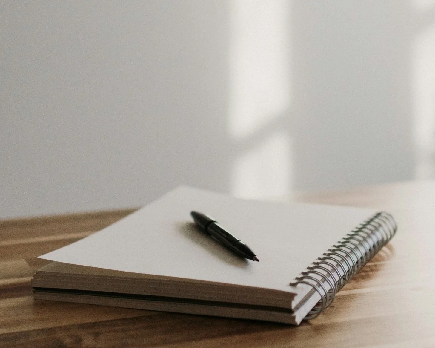 A black pen resting on an open spiral notebook on a wooden surface with a blurred background.