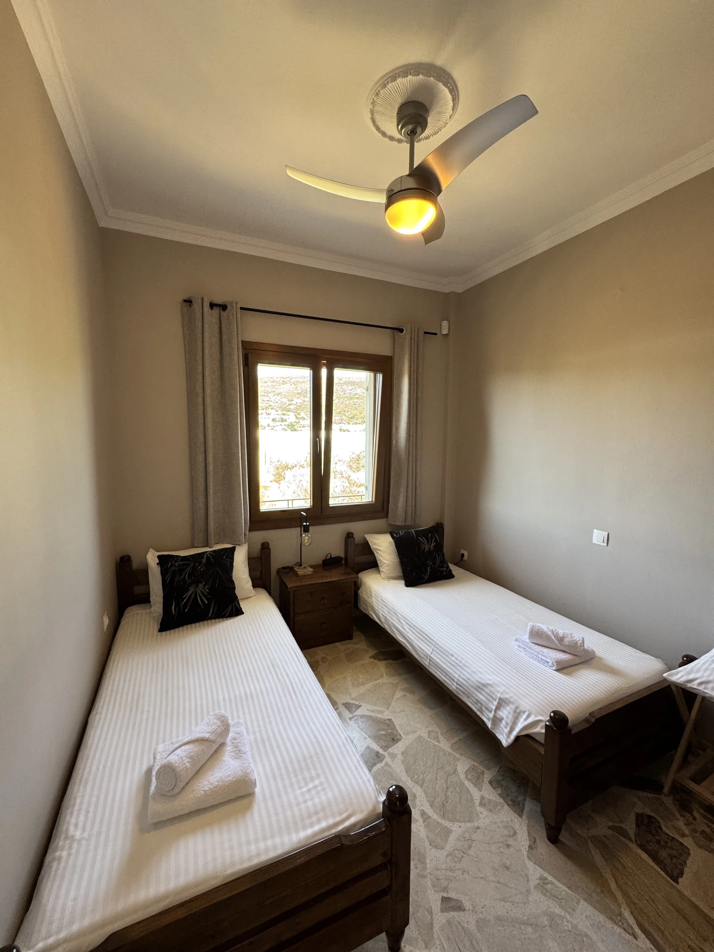 Tweedehands hotelkamer met twee eenpersoonsbedden, een klein nachtkastje met lamp, een raam met beige gordijnen, plafondventilator, en gepolijste stenen vloer.