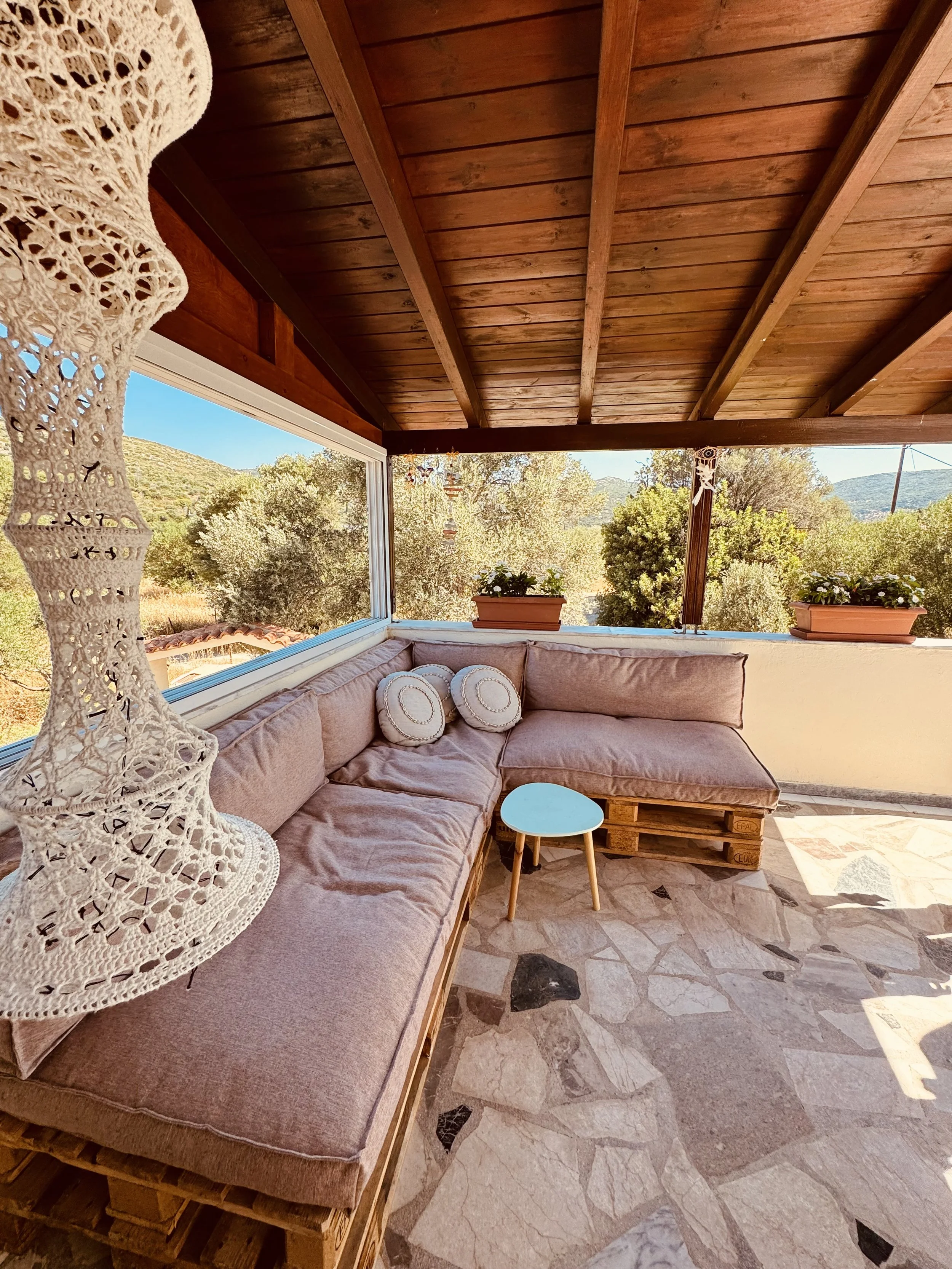 Een gezellig terras met een loungebank gemaakt van pallets, bekleed met roze kussens, en twee ronde sierkussens. Er is een kleine blauwe tafel en twee bloempotten met witte bloemen. Het terras heeft een houten plafond en uitzicht op groene bomen en h