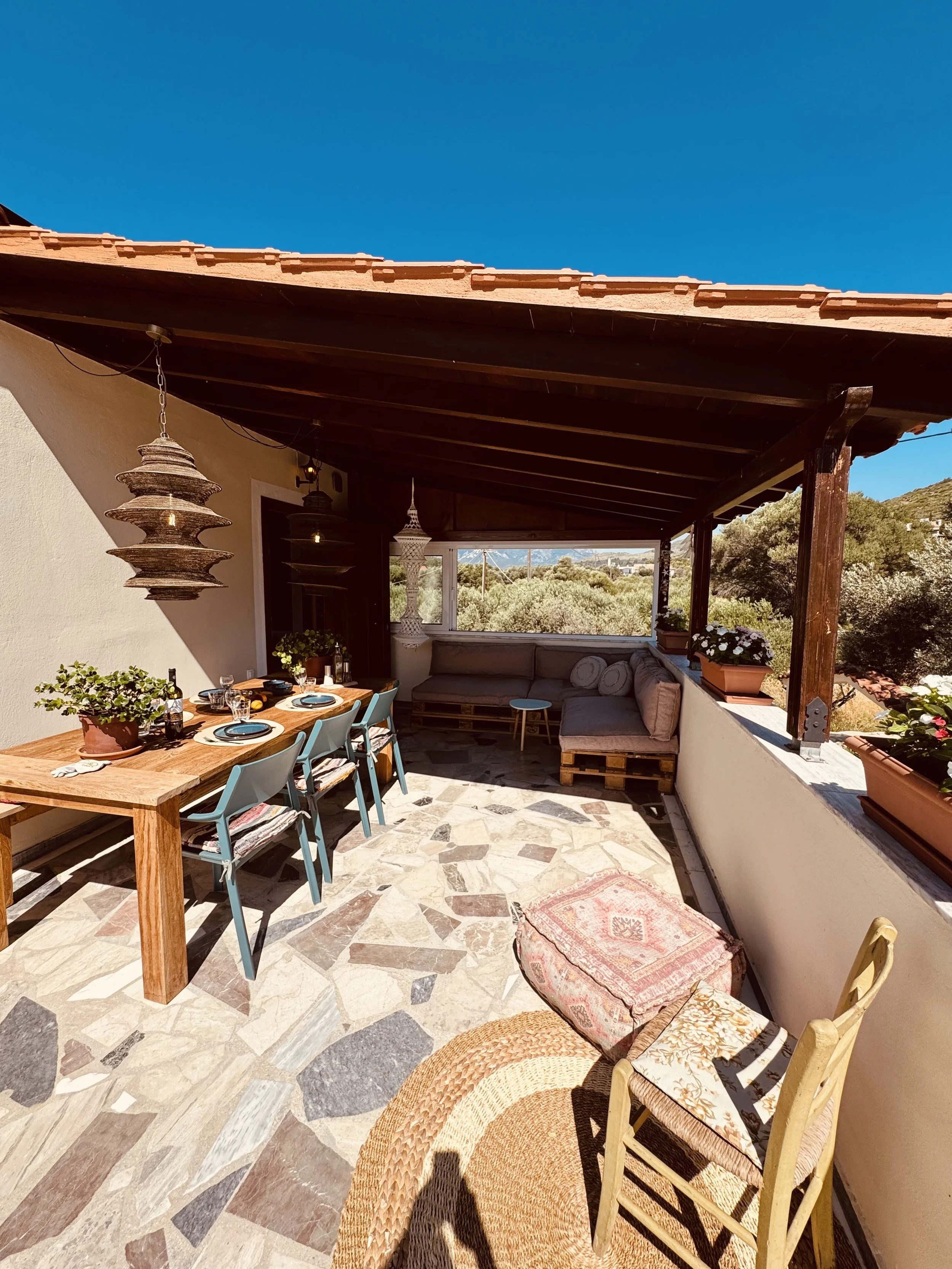 Kleine zonnige patio met eet- en zithoek, planten in potten, houten tafel met zes stoelen, banken met kussens, en decoratieve lampen, omgeven door natuurlijke omgeving en een blauwe hemel.