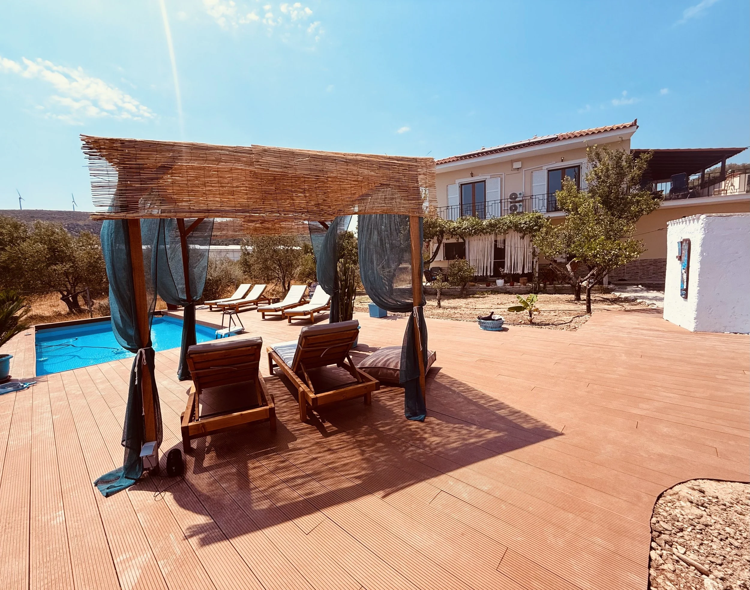 Een zonnig terras met een blauwe zwembad, houten loungebanken met kussens, een overkapping met gordijnen, een huis met balkon en tuin met bomen en planten.