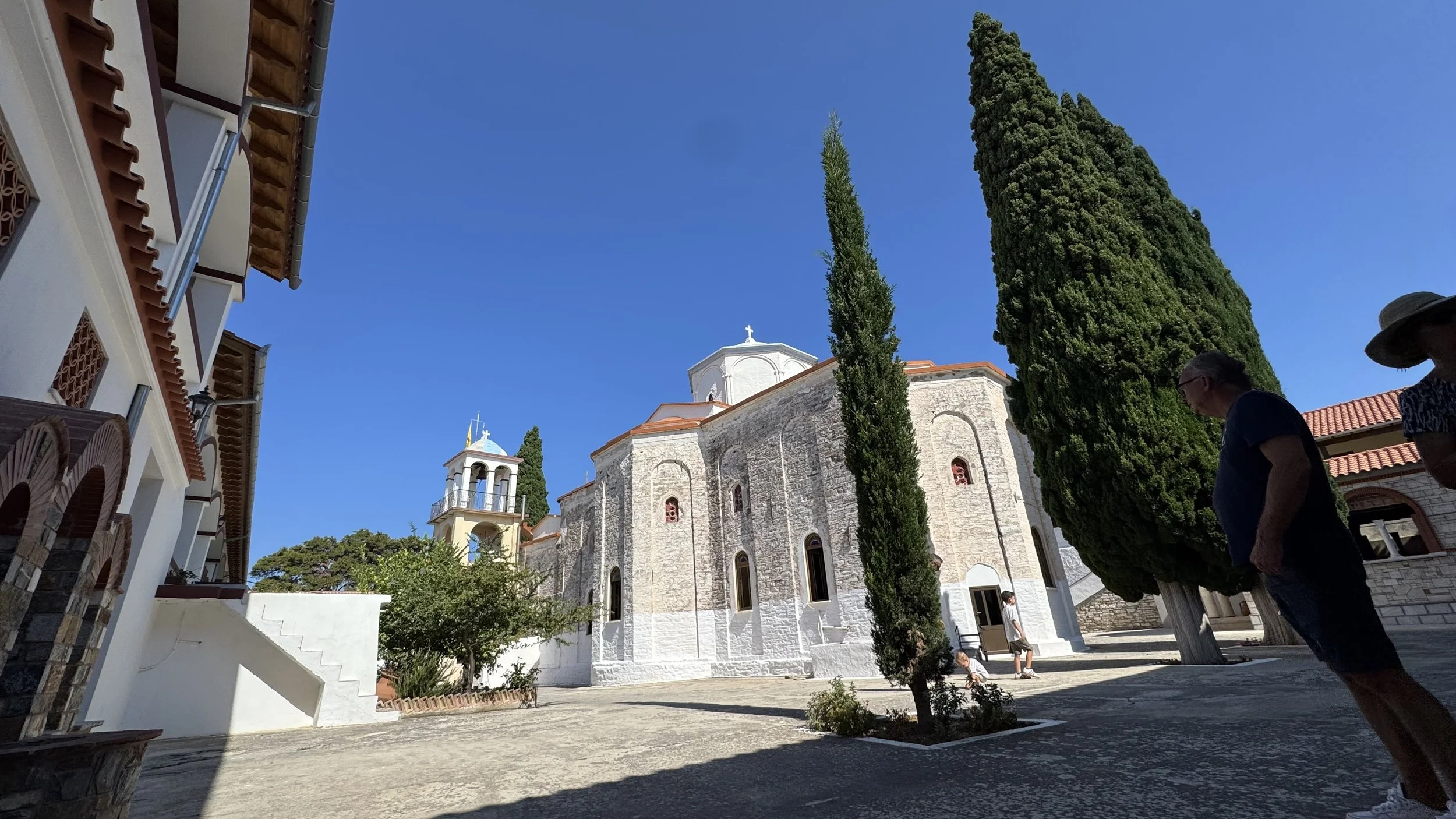 Een wit kerkje met een kleine toren en een kruis op het dak, omringd door groene cypressen en andere bomen, onder een heldere blauwe hemel. Mensen wandelen op het plein voor het kerkje.
