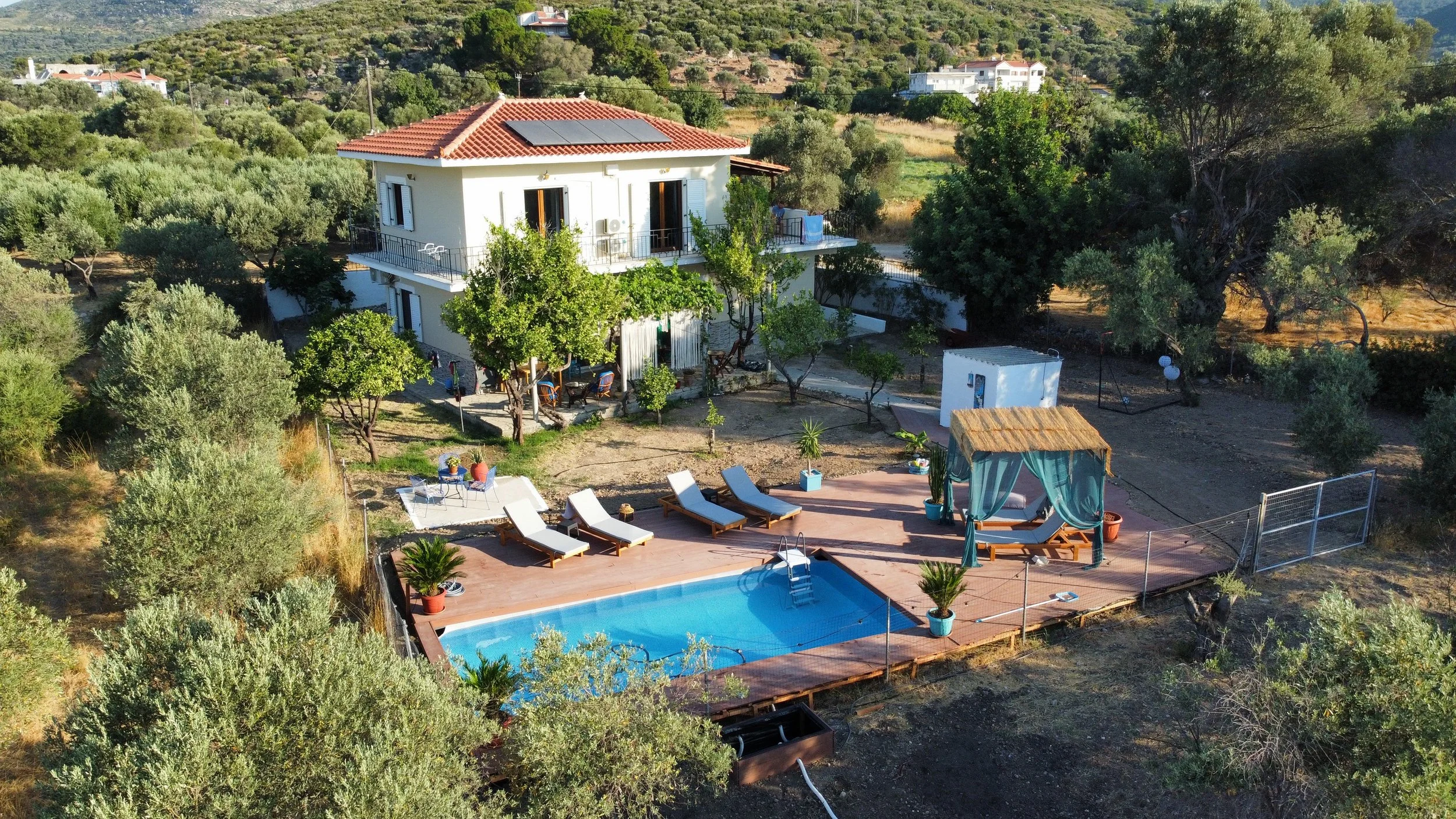 Een wit twee verdiepingen huis met een rood dak, omgeven door groene bomen en een tuin met een zwembad, ligstoelen, een schommel en een terras, gelegen in een landelijke omgeving.