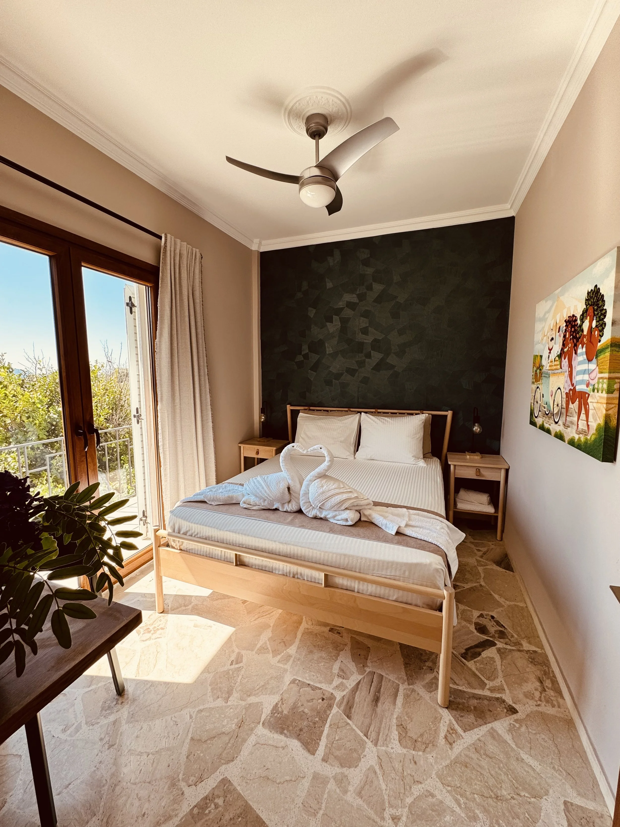 Hotelkamer met tweepersoonsbed, twee handdoekendozen in de vorm van twee zwangeren, uitzicht op balkon, plafondventilator en kleurrijk schilderij aan de muur.