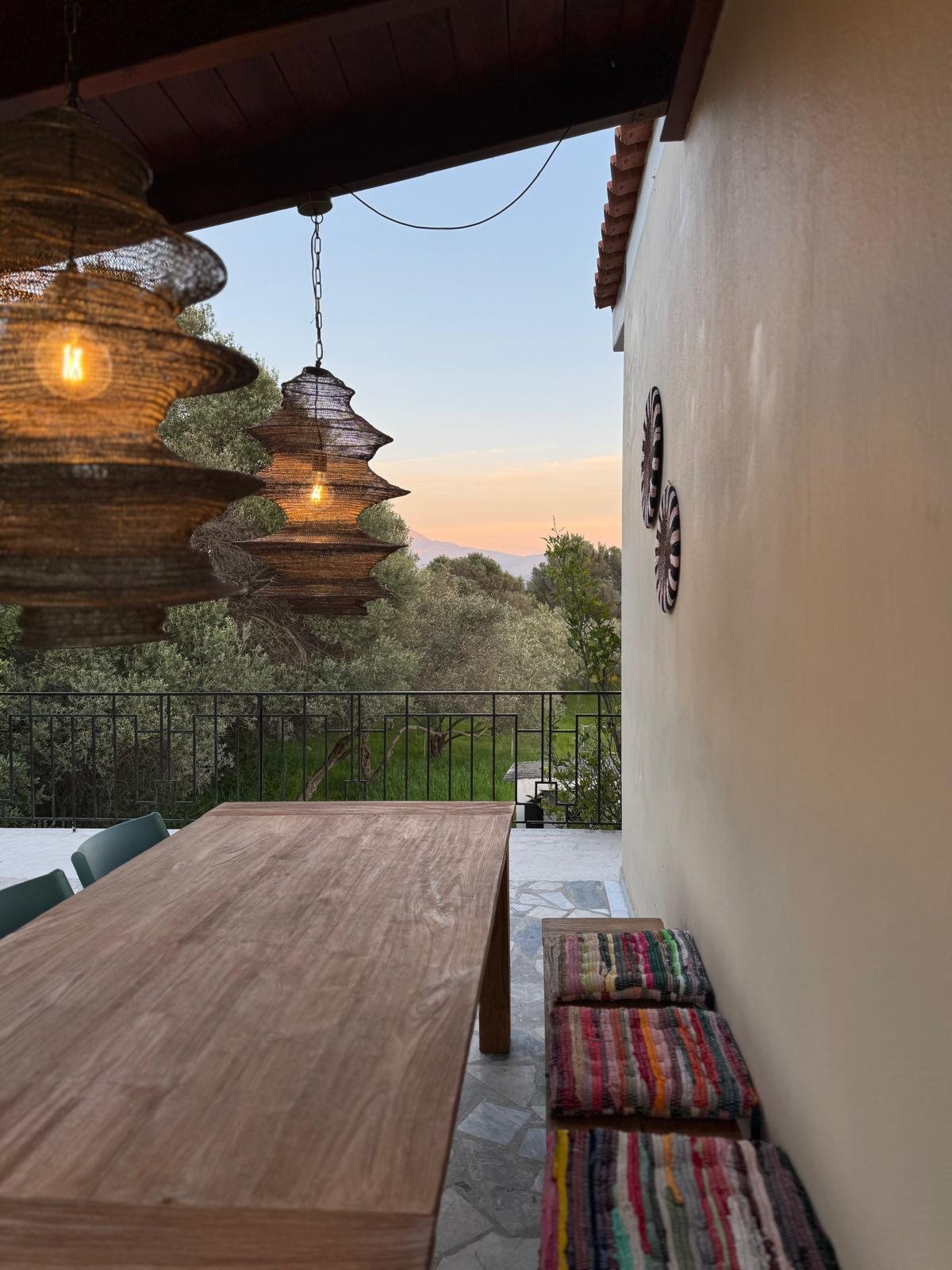Terras buitenruimte met houten tafel, stoelen, hanglampen en uitzicht op veel groen, natuur en zonsondergang.
