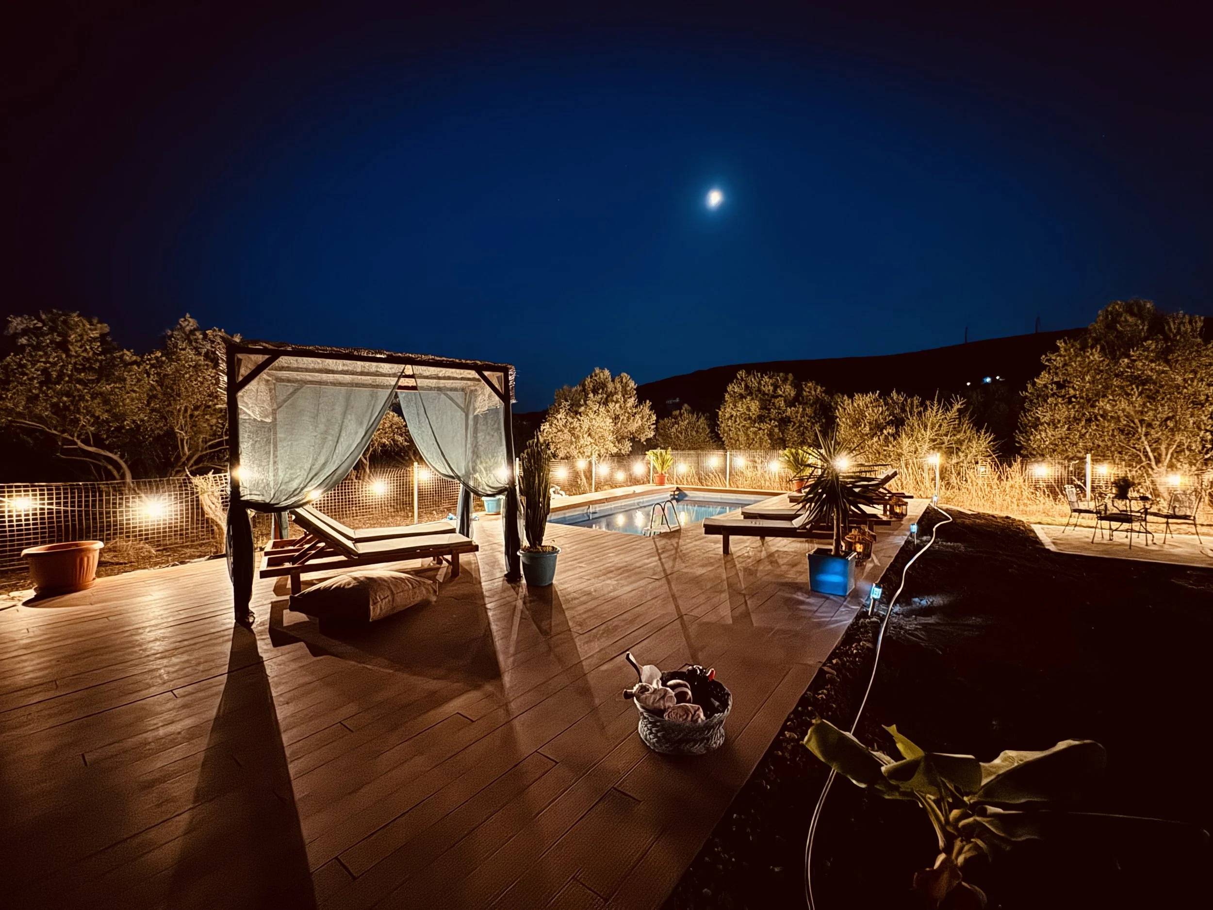 Een verlichte pool in een tuin 's avonds, met een houten terras, ligbedden, een bed met gordijnen, tuinmeubelen, tuinplanten en een hek, onder een heldere, maanverlichte hemel.