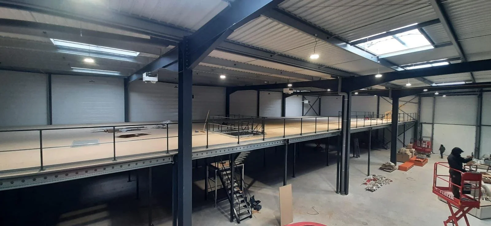 Vue intérieure d'une usine en construction avec un niveau supérieur en bois, des employés travaillant et des équipements industriels.