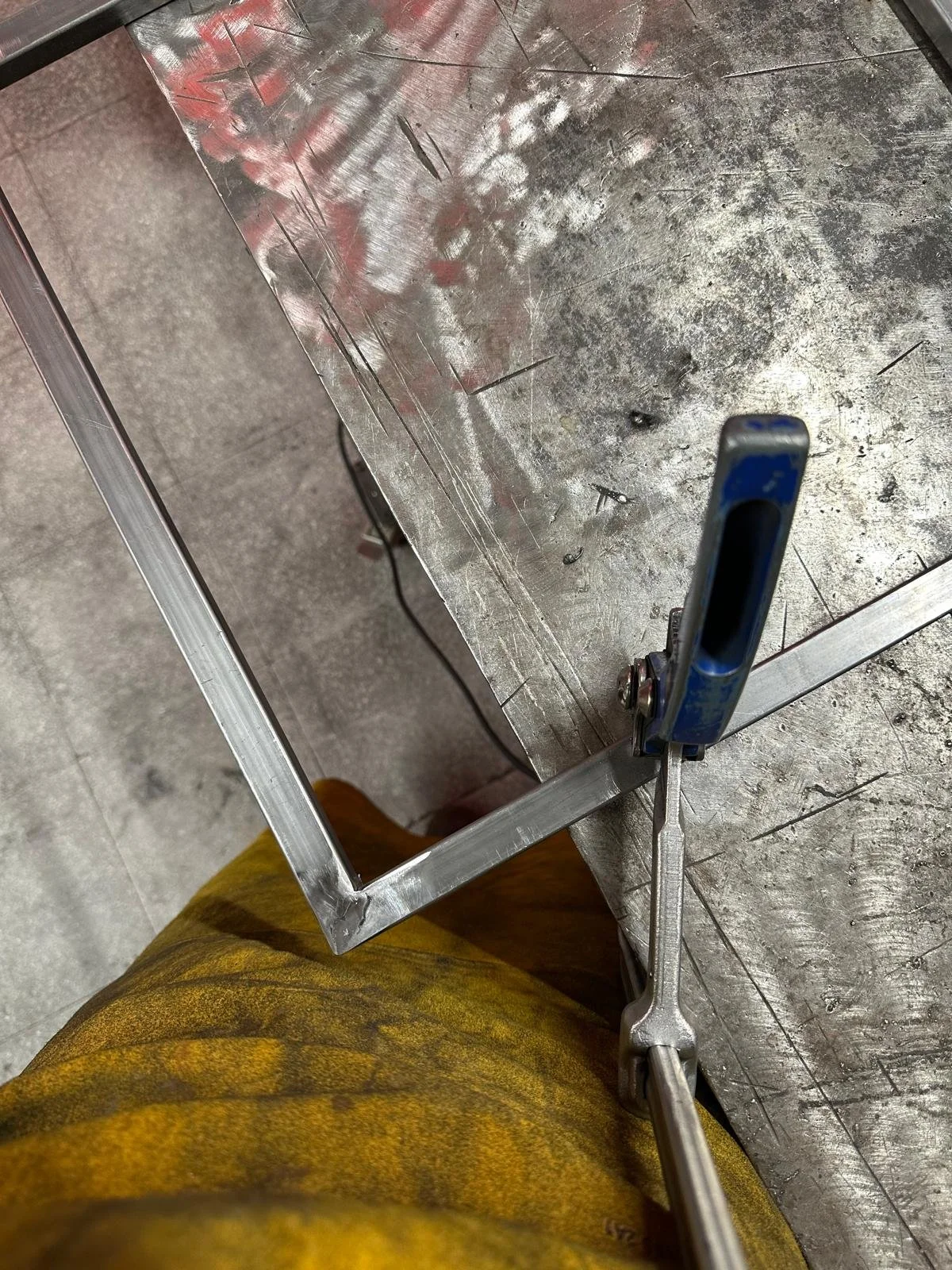 Imagen desde arriba de una mesa de trabajo de metal, con marcas y rayaduras, y una herramienta de apriete en la mano de una persona con pantalones amarillos. La mesa tiene un marco metálico y en el fondo se ve parte del piso.