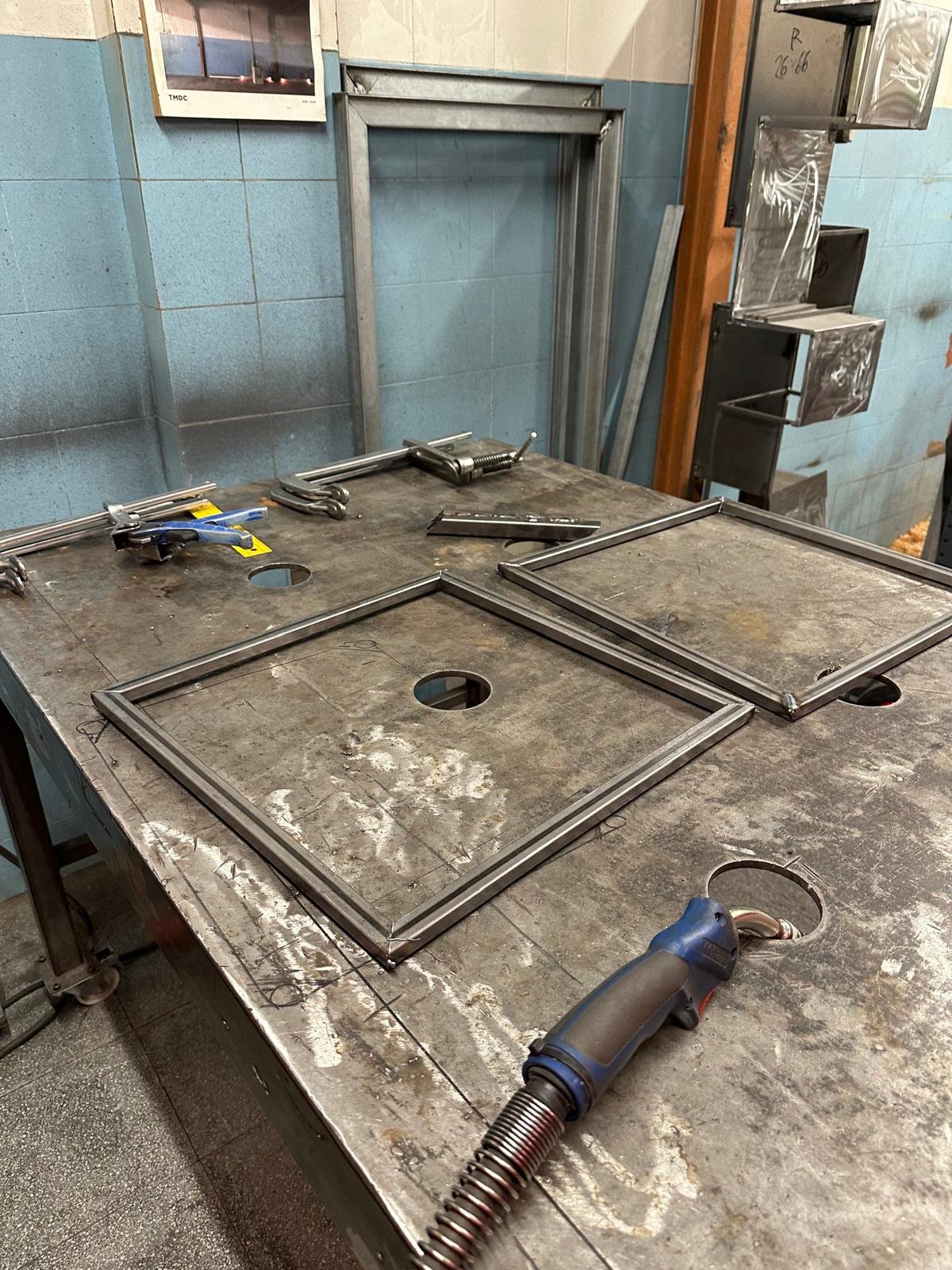 Mesa de trabajo en una herrería con marcos de metal y herramientas, y un equipo de soldadura con tubo flexible.