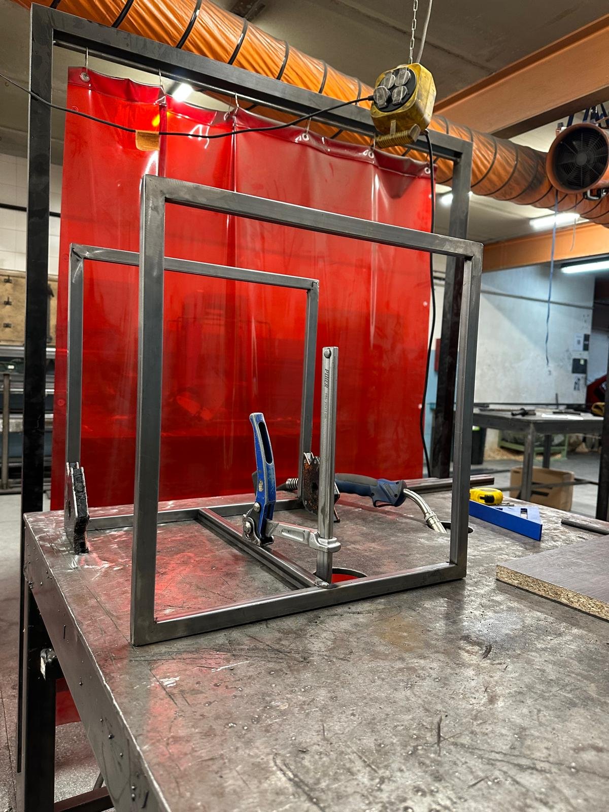 Una estructura de marco de aluminio en proceso de construcción en un taller de metalurgia, con herramientas como una pinza y una lijadora en una mesa de trabajo, fondo con cortinas de protección roja y conductos de ventilación de color naranja.