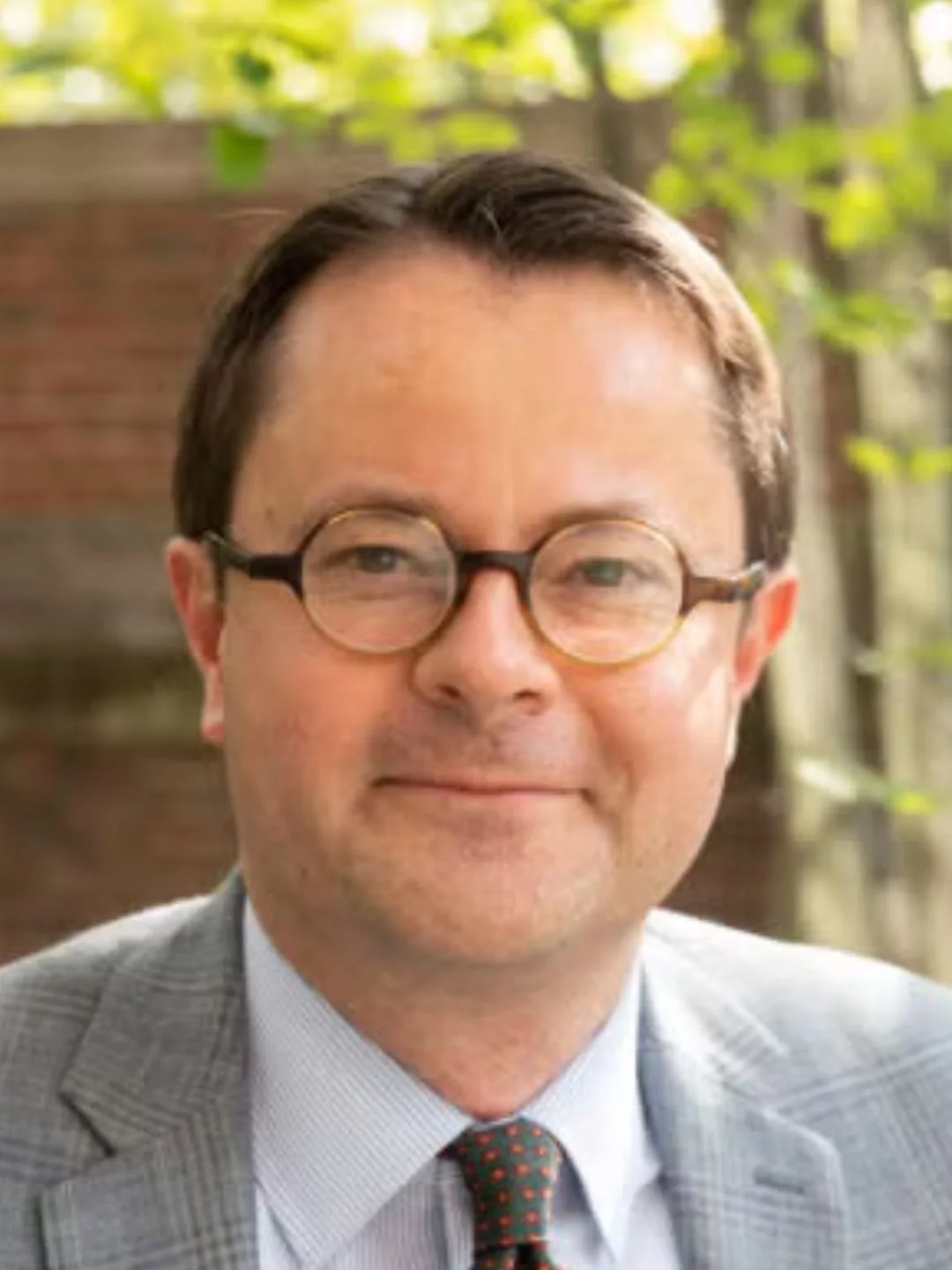 A person wearing round glasses and a suit, smiling slightly against a blurred background.