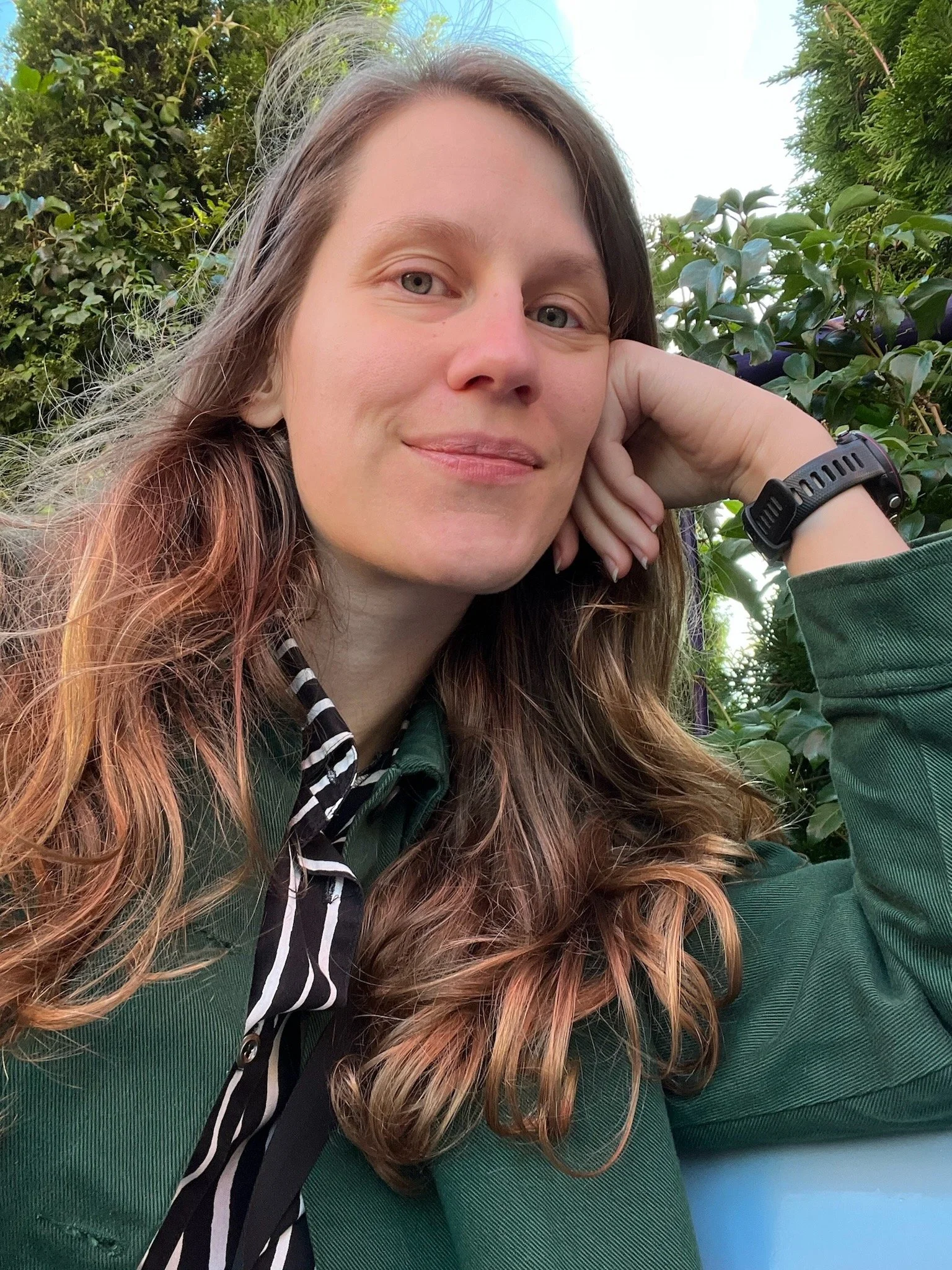 Heini, a white woman with long wavy brown hair wearing a green jacket and resting her head on her hand. She is outdoors, smiling and looking straight toward the camera.