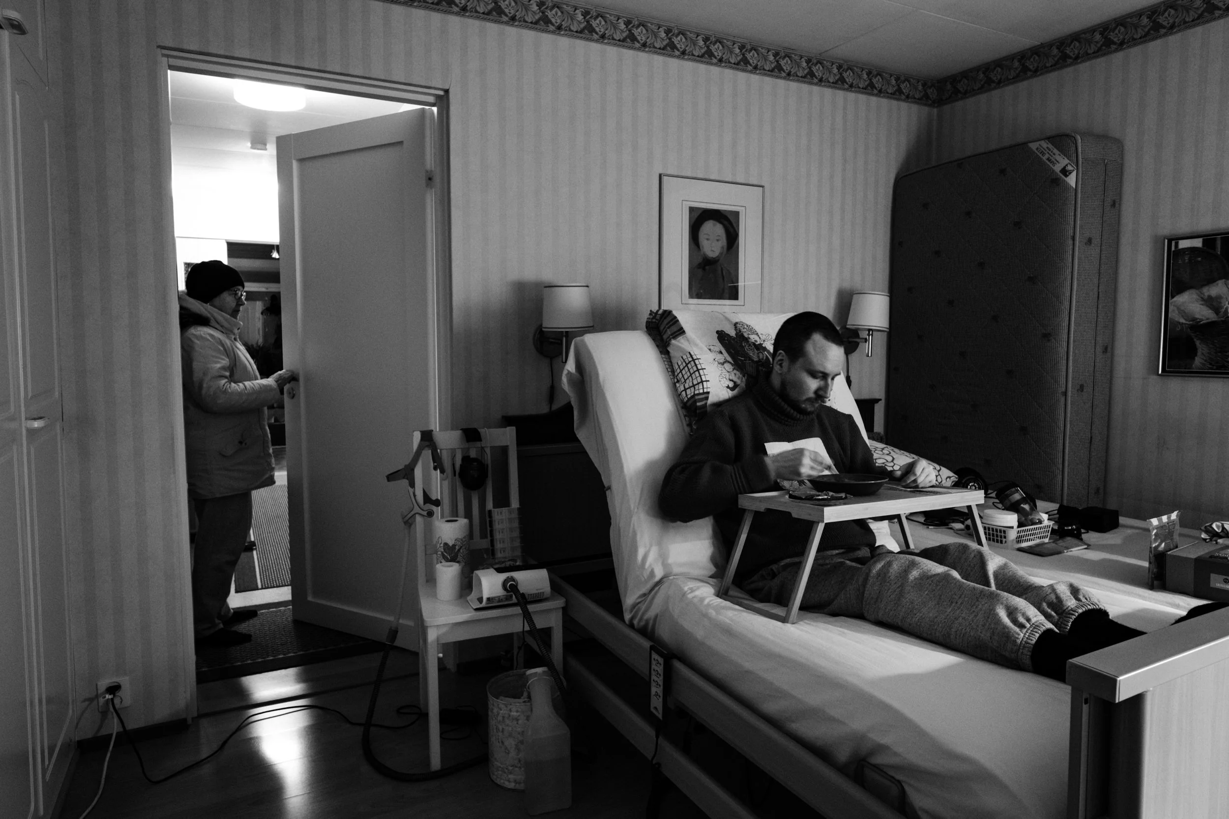 A black-and-white photo of Niko sitting upright in bed, eating from a tray table while wearing a sweater. Päivi, in a winter coat, stands in the open doorway looking in. A CPAP machine and medical equipment are beside the bed.
