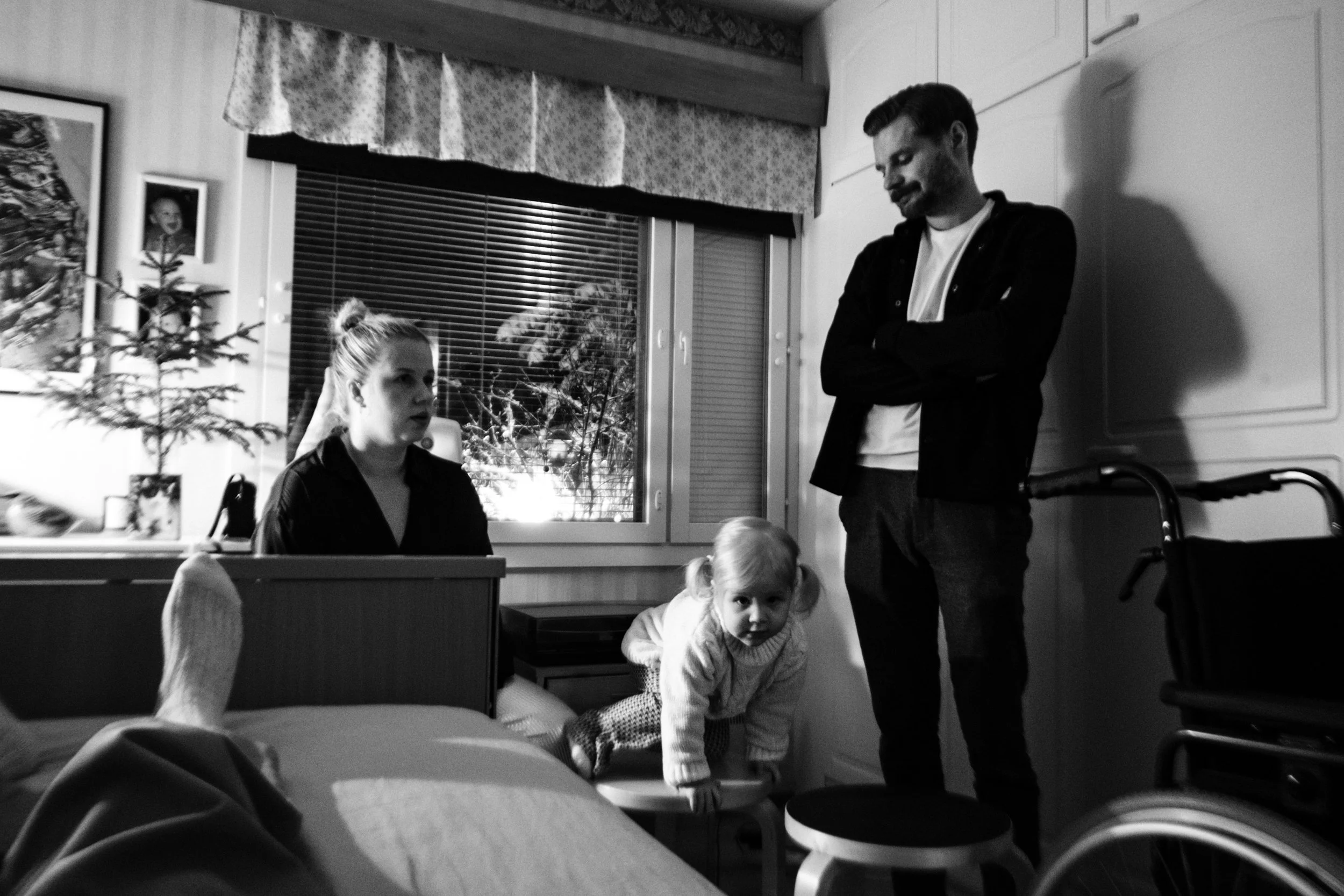 A black-and-white photo from the perspective of Niko lying in bed. A woman with a bun sits near the bed. A bearded man stands with arms crossed. A young child with pigtails climbs a stool between them. A wheelchair is in the background.