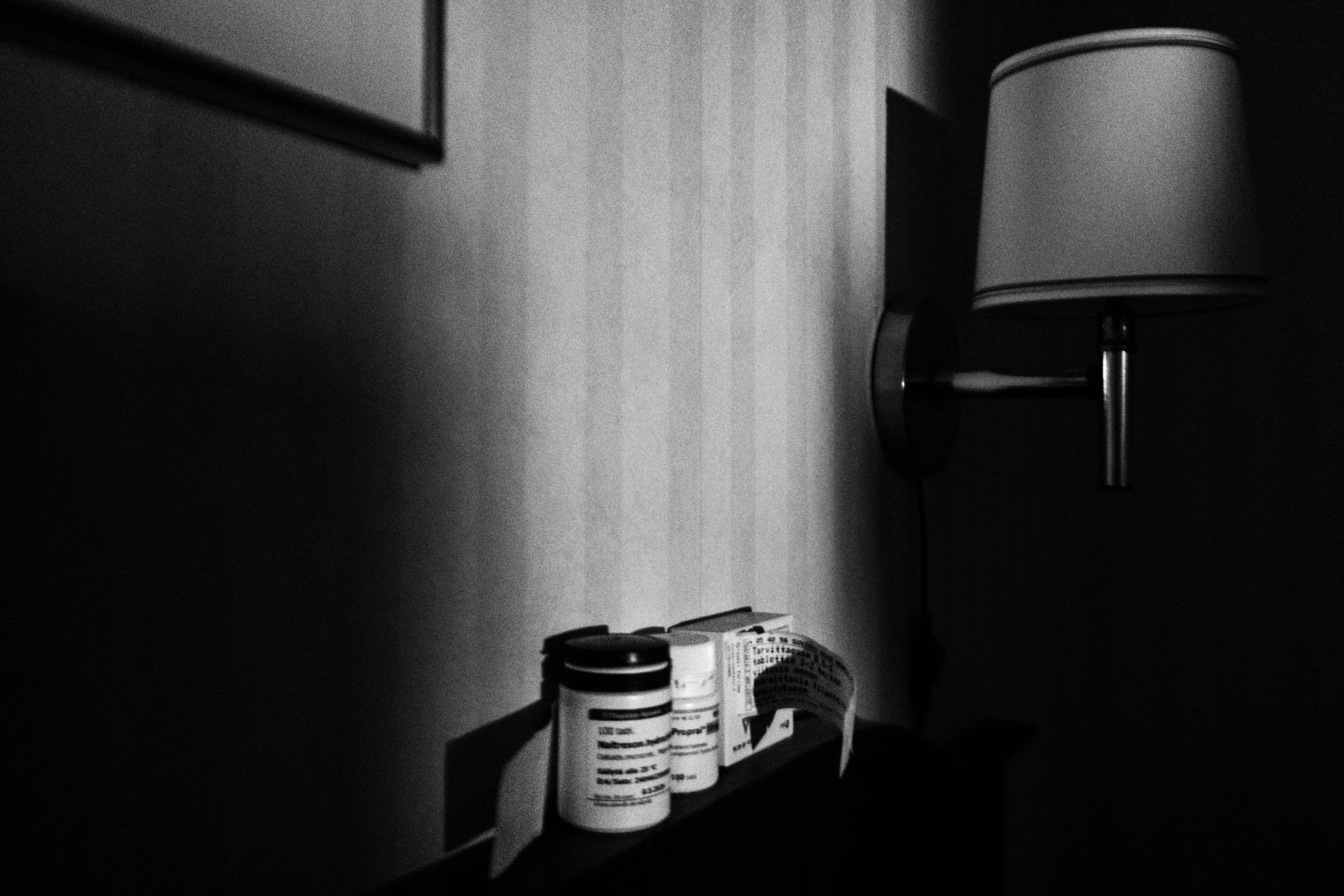 A black-and-white photo of three medication bottles on top of a bedframe, the light illuminates the bottles in an otherwise dark bedroom. A wall lamp and a frame are also part of the picture.