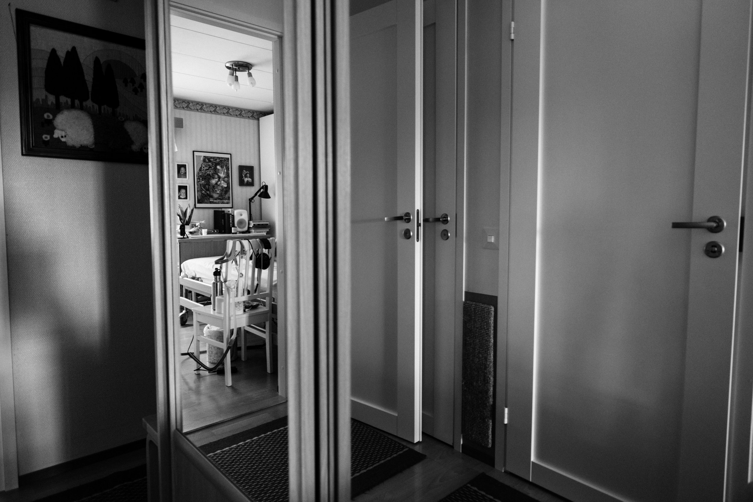 A black-and-white photo of a hallway’s cabinet with mirror doors. The mirrors reflect a bedroom seen from a doorway and multiple doors, which makes the image appear distorted and abstract.