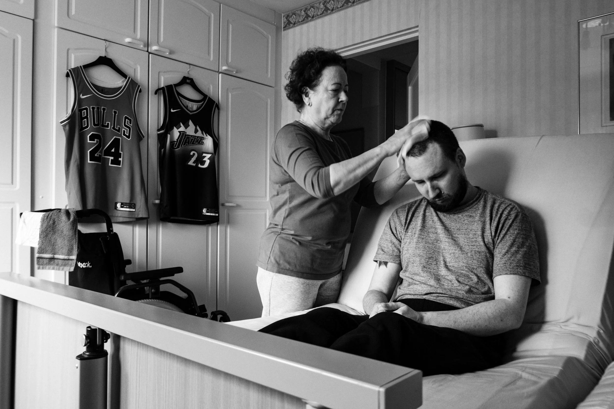 A black-and-white photo of Päivi wiping Niko’s head as he sits with a solemn expression in a home care setting. He wears a gray t-shirt. In the background, Chicago Bulls and Utah Jazz jerseys hang on a wardrobe, and a wheelchair is visible.