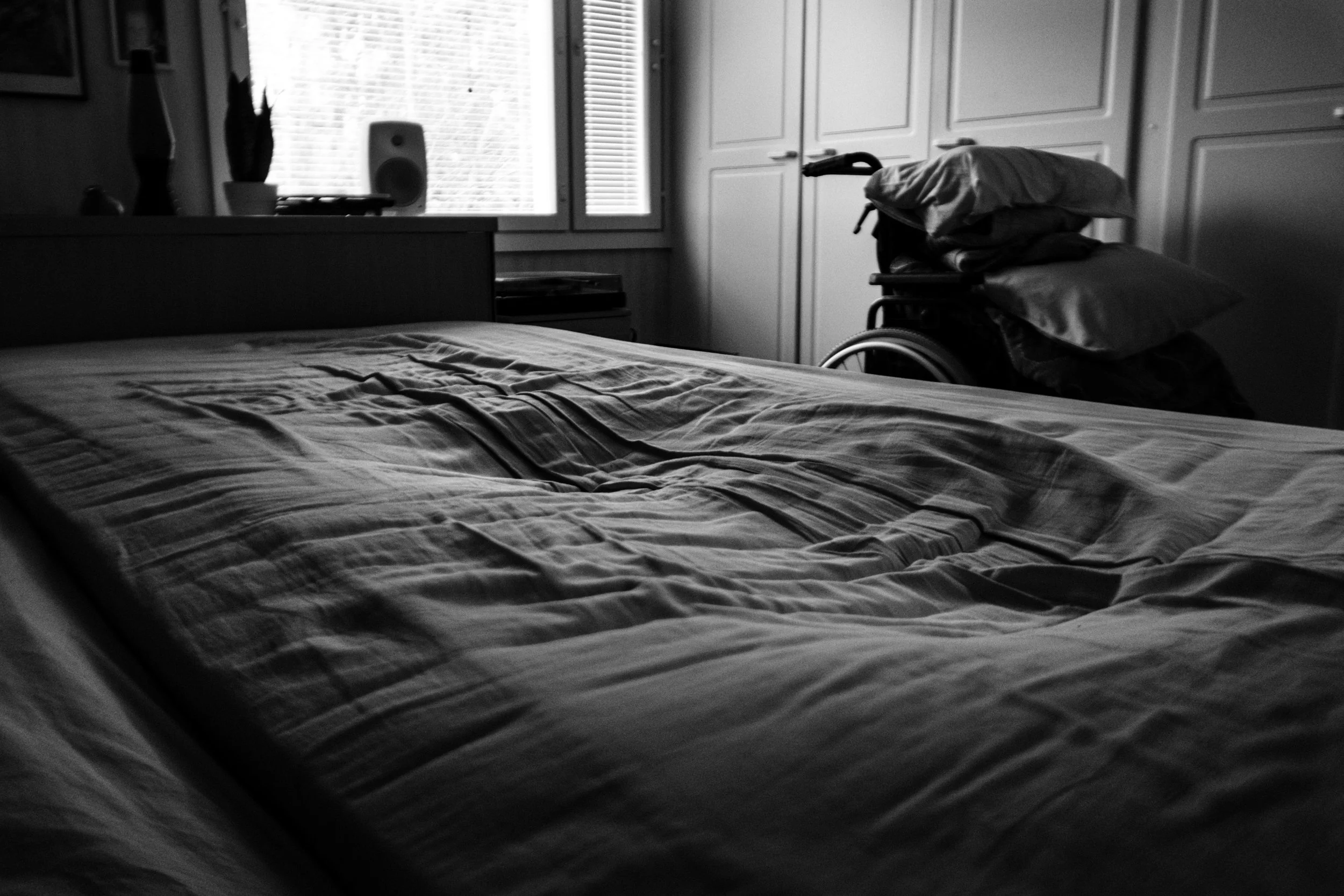 A black-and-white photo of an empty bed, which shows the dip of a human silhouette. A wheelchair, personal belongings, and a window are seen in the background.