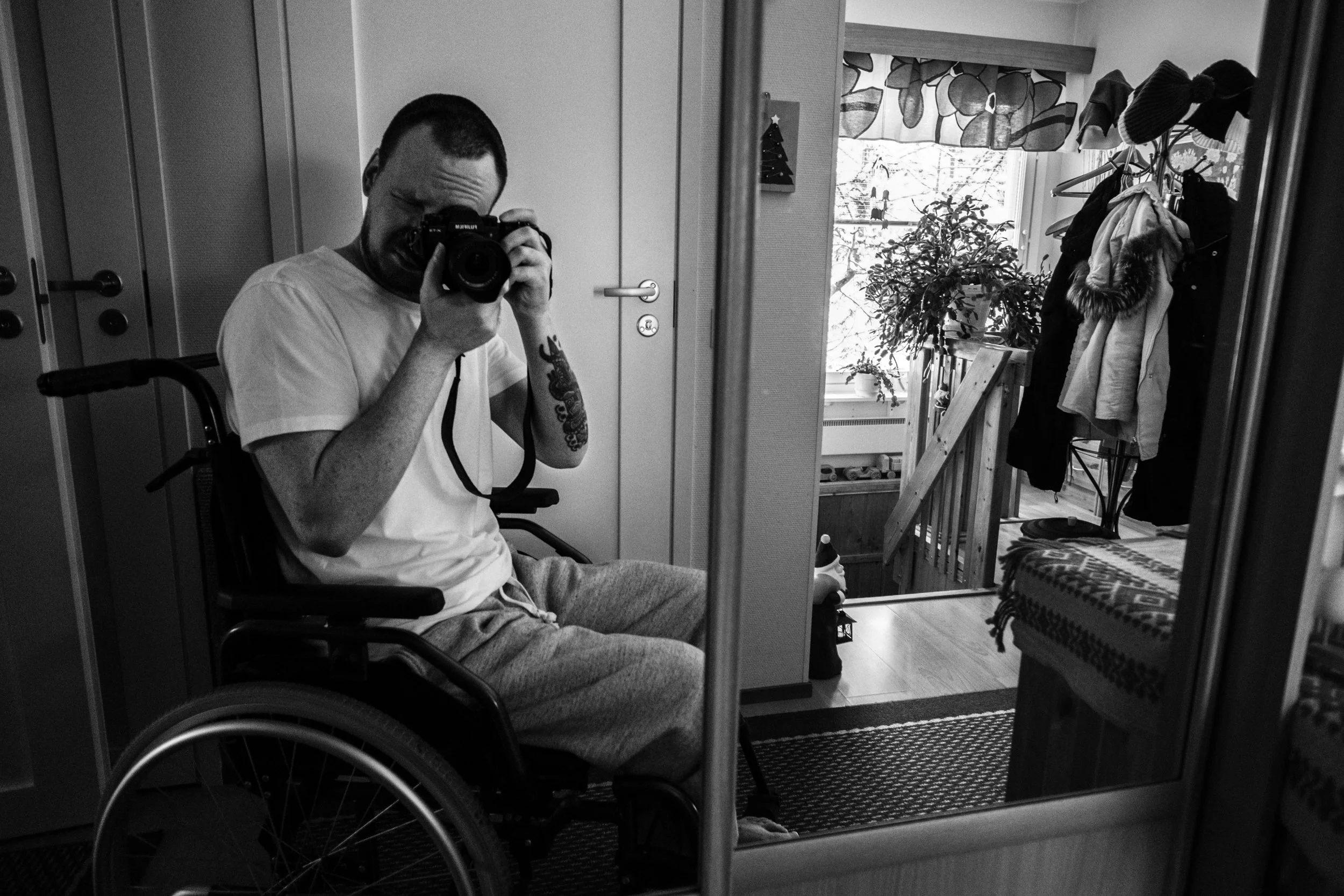 A black-and-white photo of Niko, with short hair and a beard, wearing a white t-shirt and grey sweatpants, sitting in a wheelchair. He takes a self-portrait through a cabinet’s mirrored door. A rack of clothes and a window are in the background.