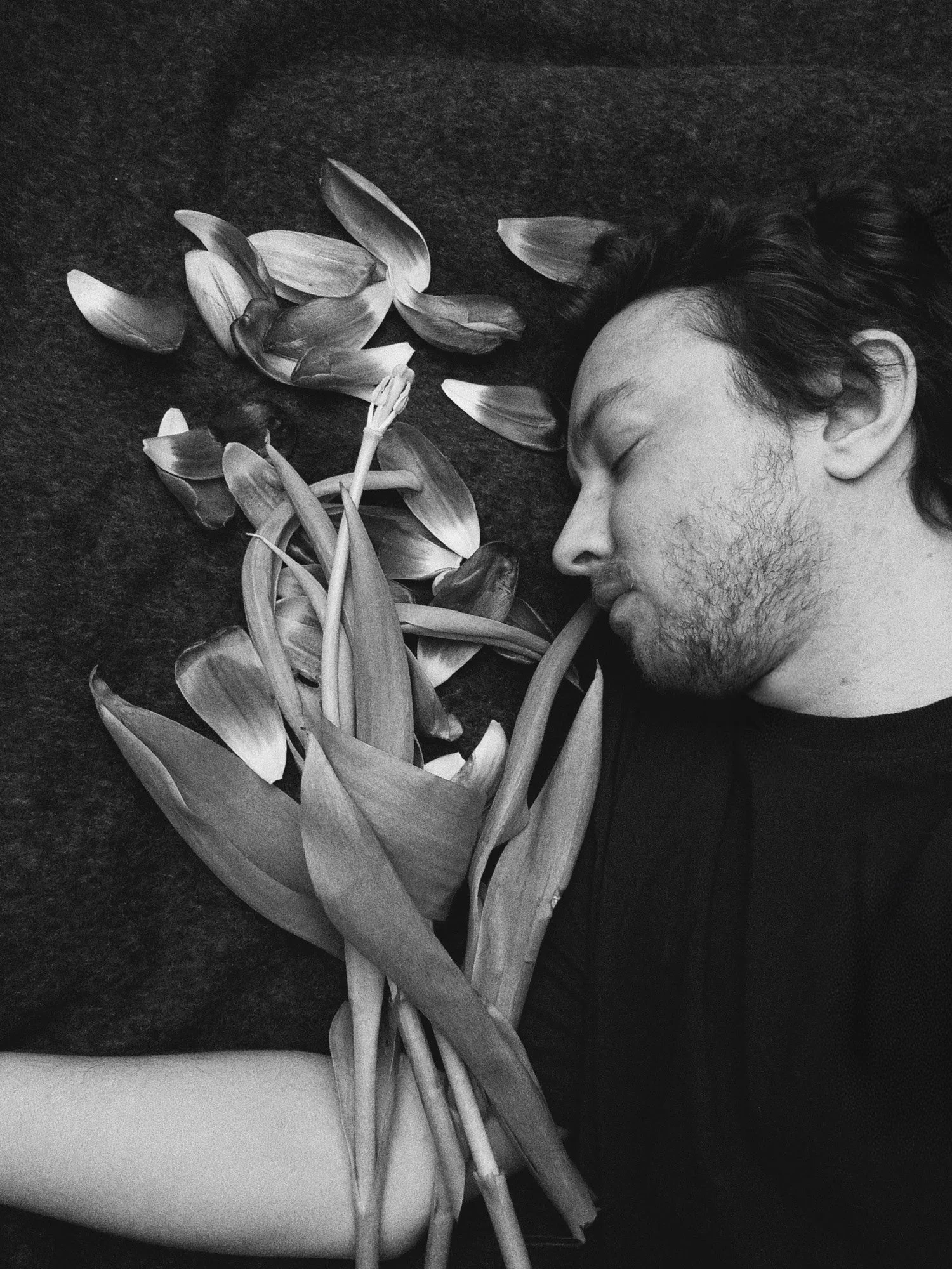 A black-and-white photograph of a man with his eyes closed lying down on a dark blanket next to a bouquet of dead flowers.