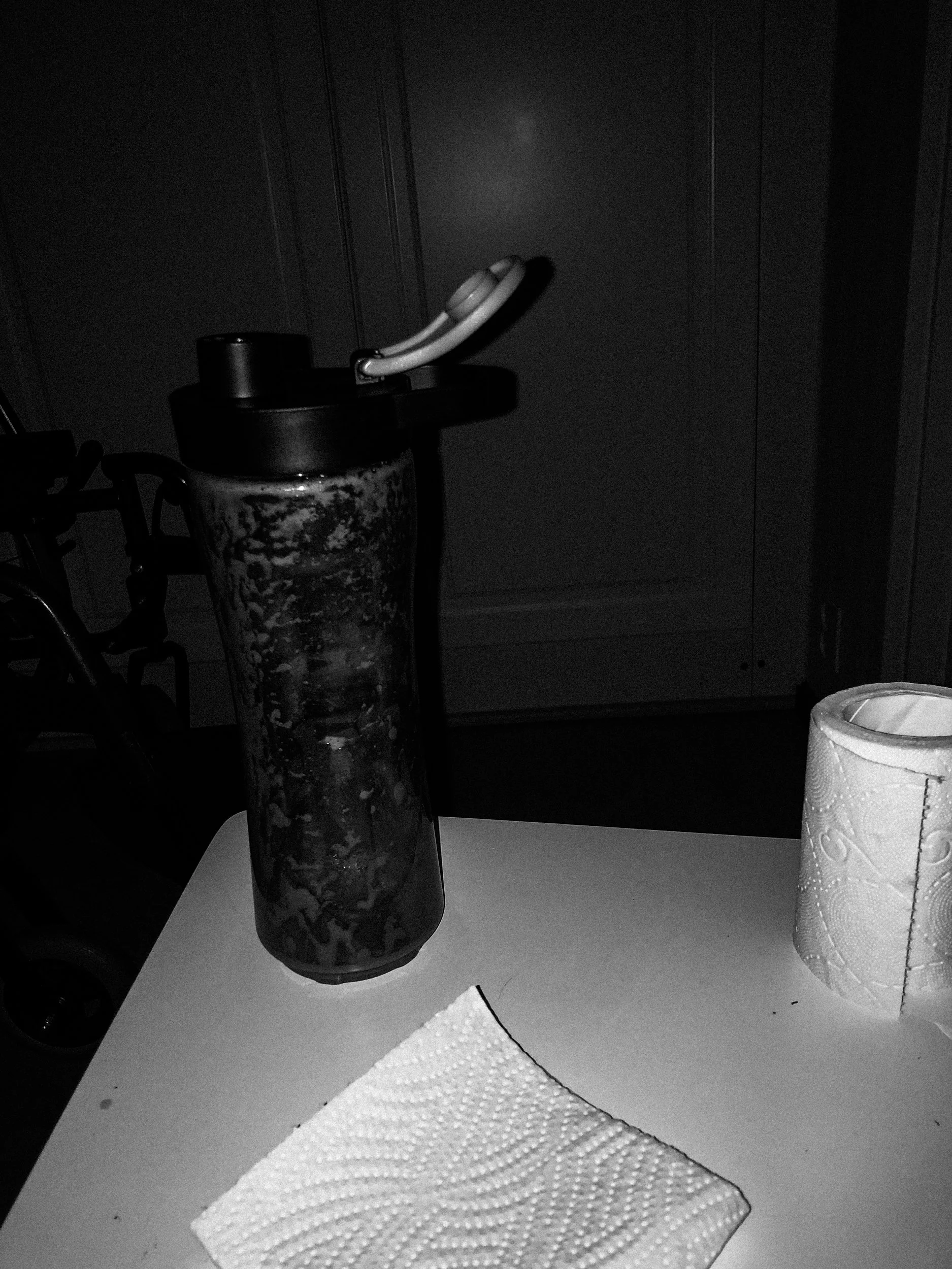 A black-and-white photo of a bedside chair that has a drinking bottle, a piece of paper and a roll of toilet paper on it, all lit up by the camera’s flash in a dark bedroom.
