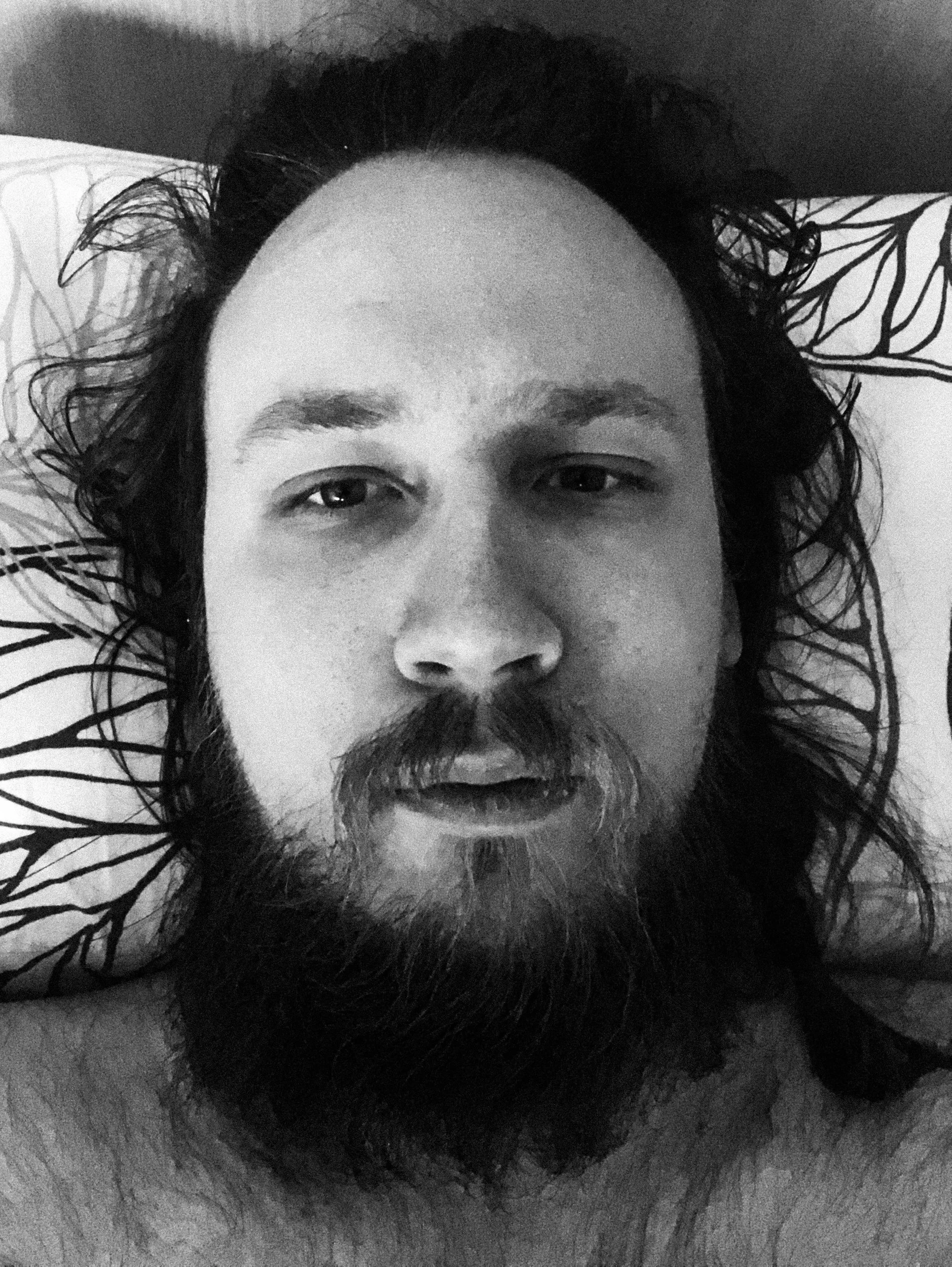 A black-and-white selfie photo of Niko; he’s looking directly at the camera while lying in bed; he has a messy beard and long hair. The image has more brightness in it.