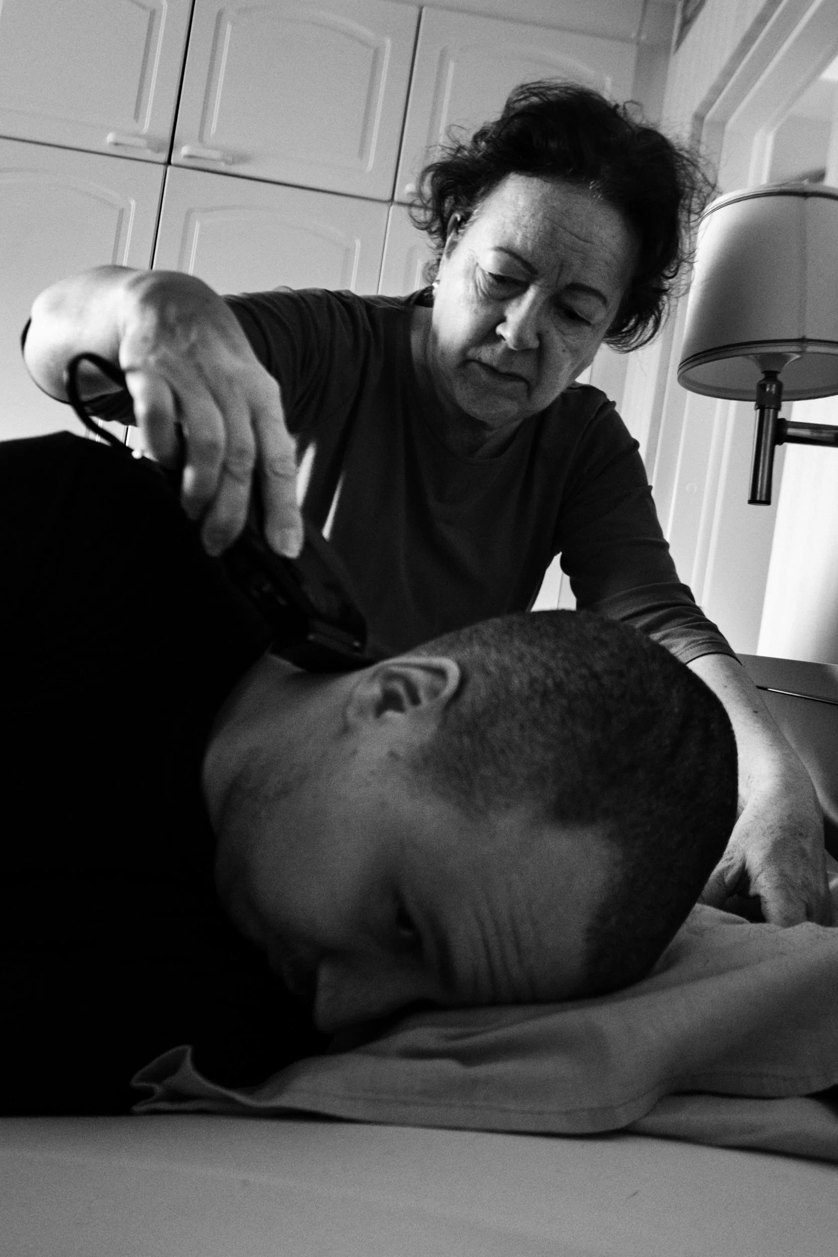 A black-and-white photo of Päivi giving a haircut to Niko, who is lying face down on a bed. She uses clippers with a focused expression. The bedroom has cabinets in the background and a bedside lamp.