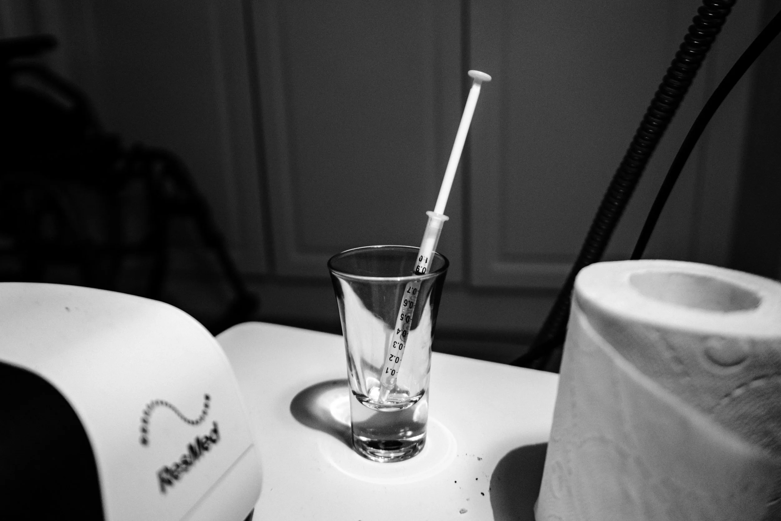 A black-and-white photo of a medication syringe full of liquid medication, placed in a shot glass. In the foreground are a roll of toilet paper and a CPAP machine.