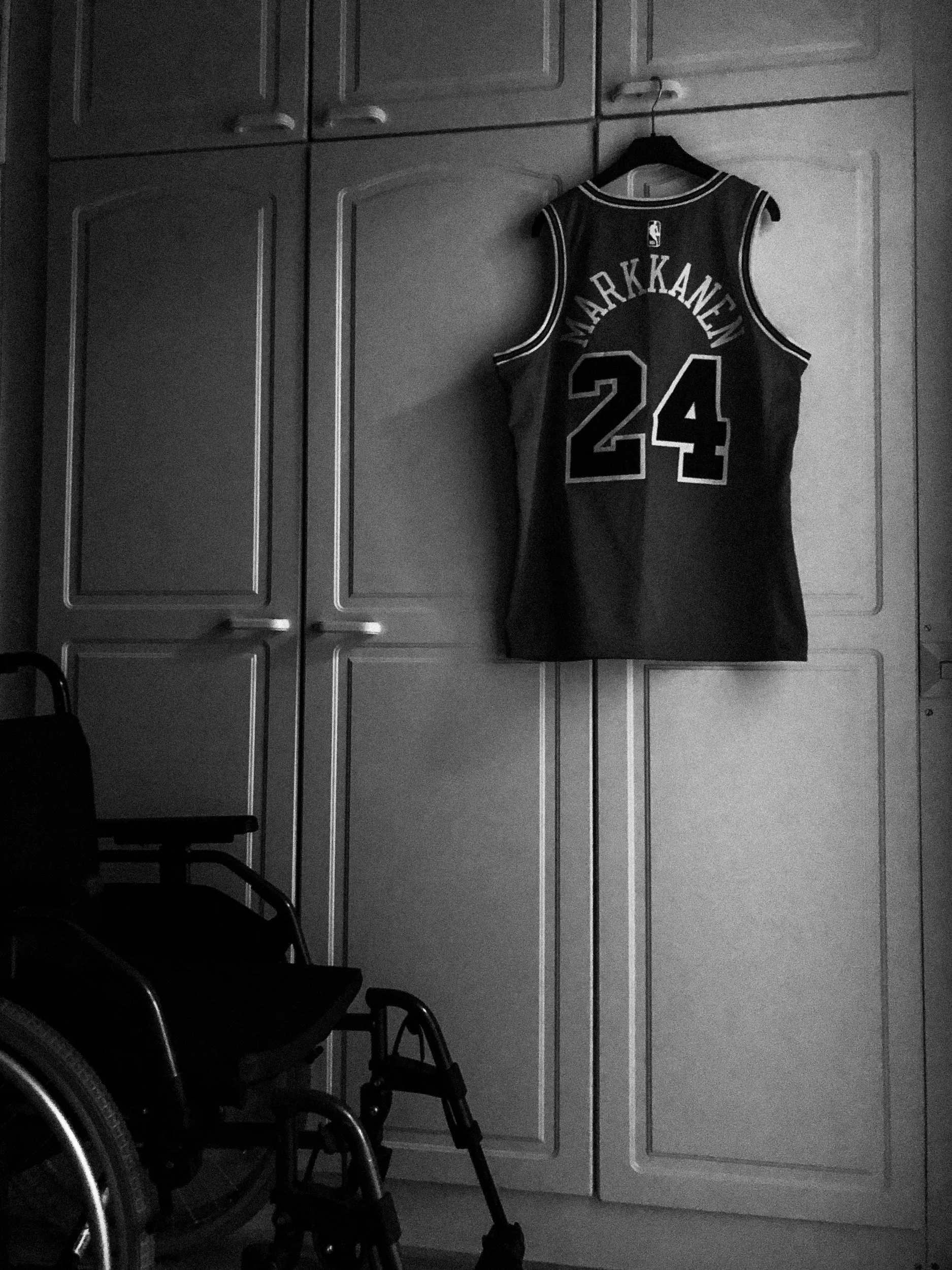 A black-and-white photo of a Chicago Bulls Markkanen jersey hanging against a white cabinet door, in a dark room. Part of a wheelchair is shown in the left corner.