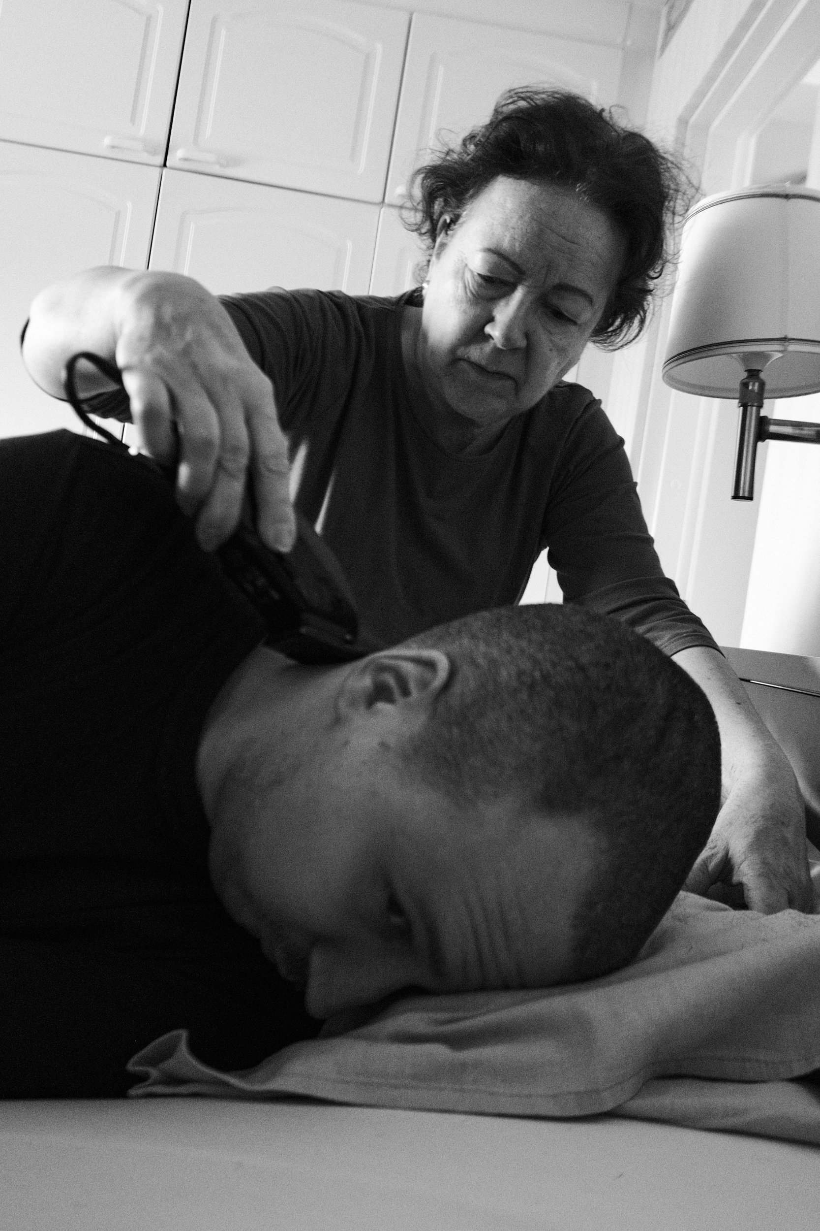 A black and white photo of Päivi giving a haircut to Niko, who is lying face down on a bed. She uses clippers with a focused expression. The bedroom has cabinets in the background and a bedside lamp.