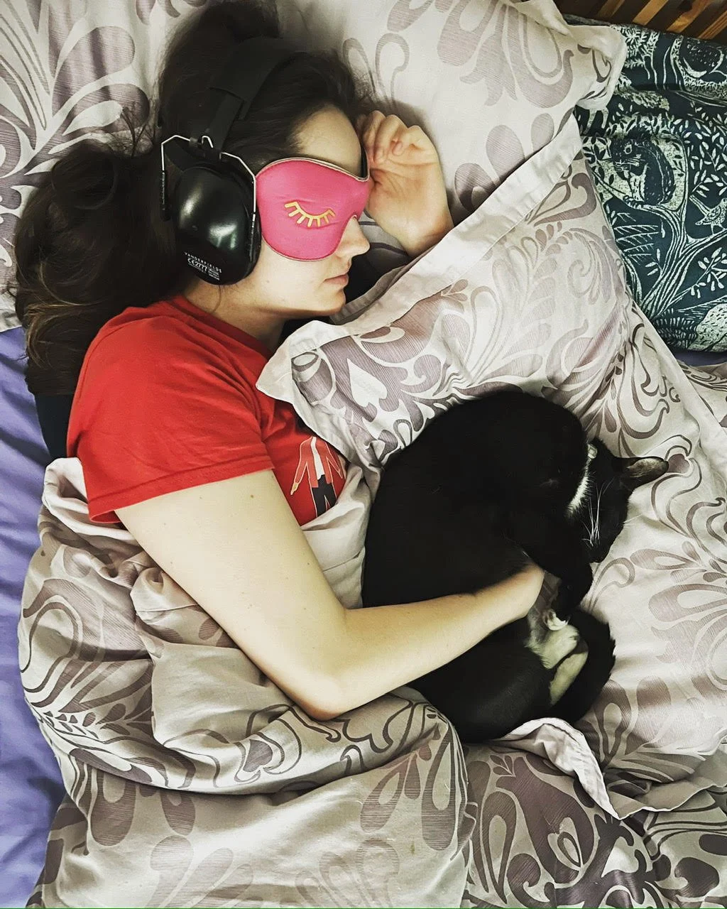 Fran, a young woman with fair skin and brown wavy hair, is wearing ear defenders and an eyemask as she lies on her side in bed. A tuxedo colored cat is curled up in her arm, matching her position.