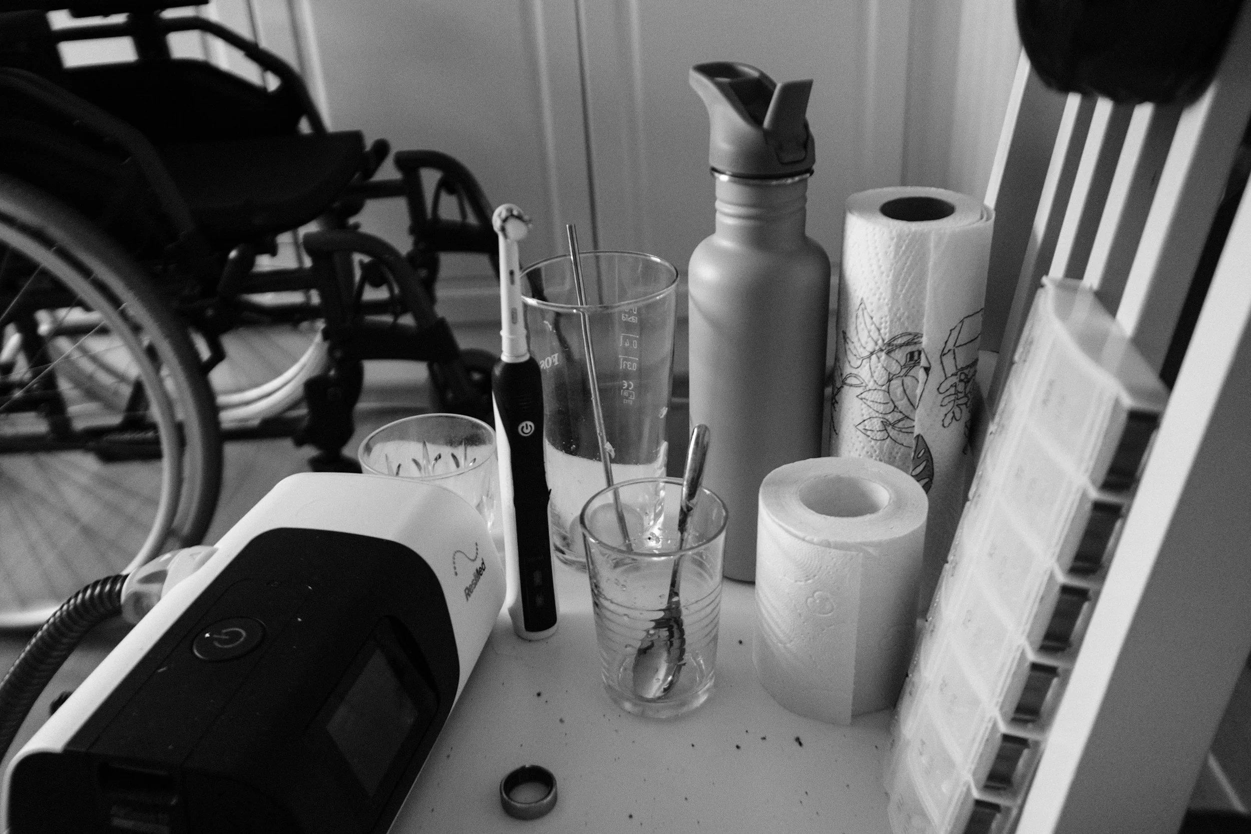 A black and white photo of a cluttered chair with an electric toothbrush, water bottle, paper towel roll, pill organiser, and other personal items. A wheelchair is in the background.