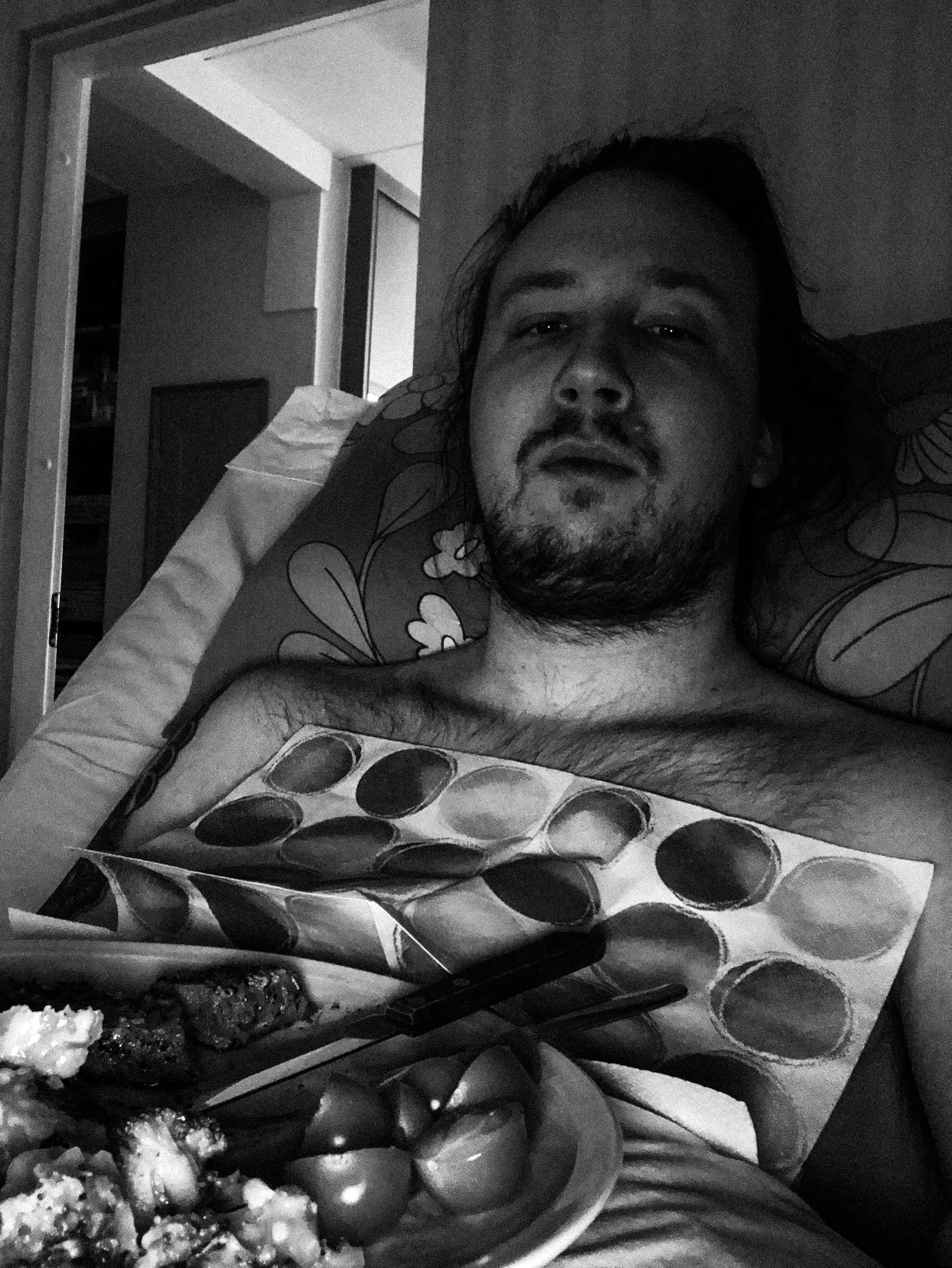 A black-and-white selfie photo of Niko in a dark bedroom, he’s looking directly at the camera while lying in bed, he has a messy short beard and long hair. He has a plate of food lying on his stomach.