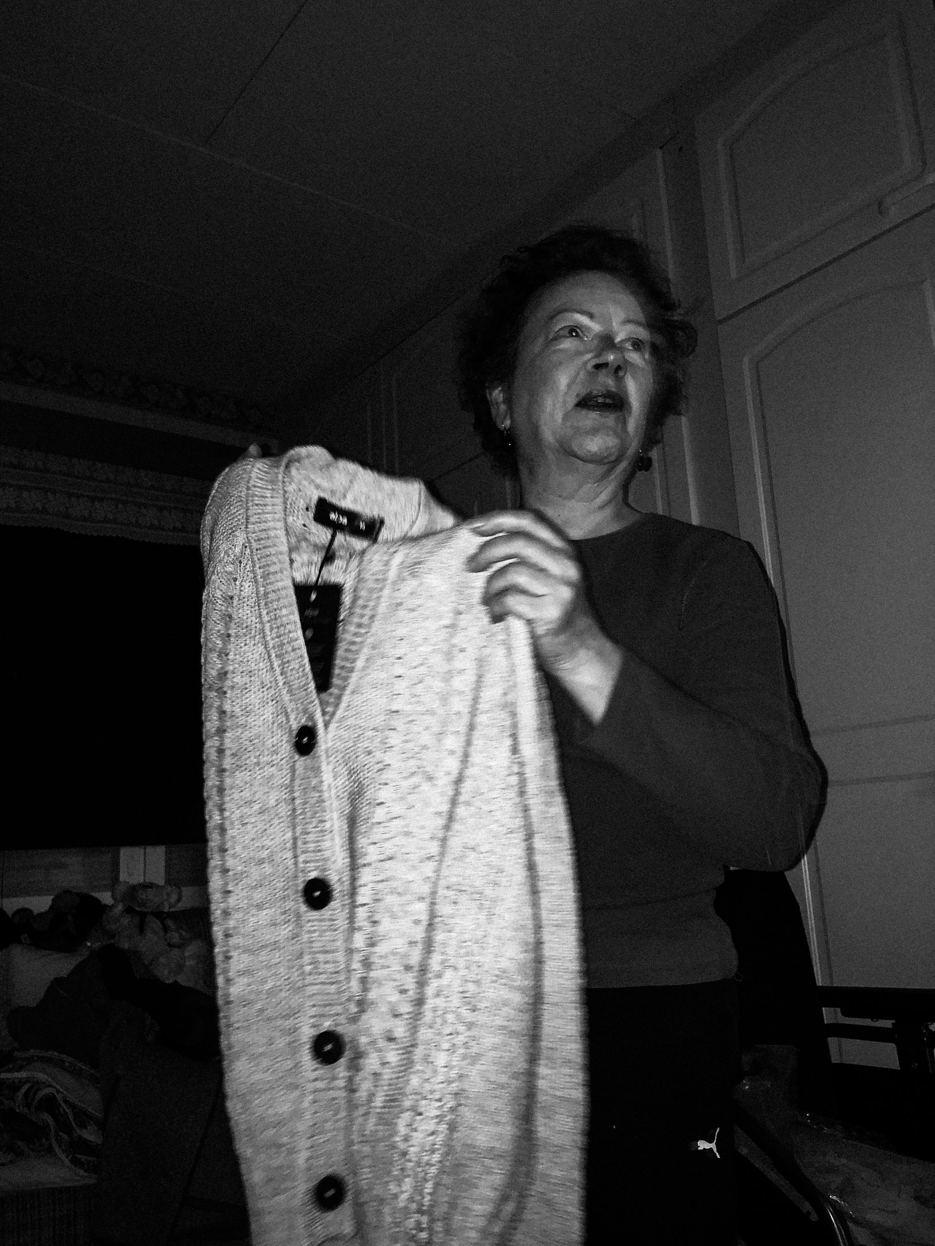 A black-and-white photo of Niko’s mother, receiving a cardigan as a gift. She is holding the cardigan in her hands. She is lit up by the camera’s flash and her face appears to be a bit blurry due to motion.
